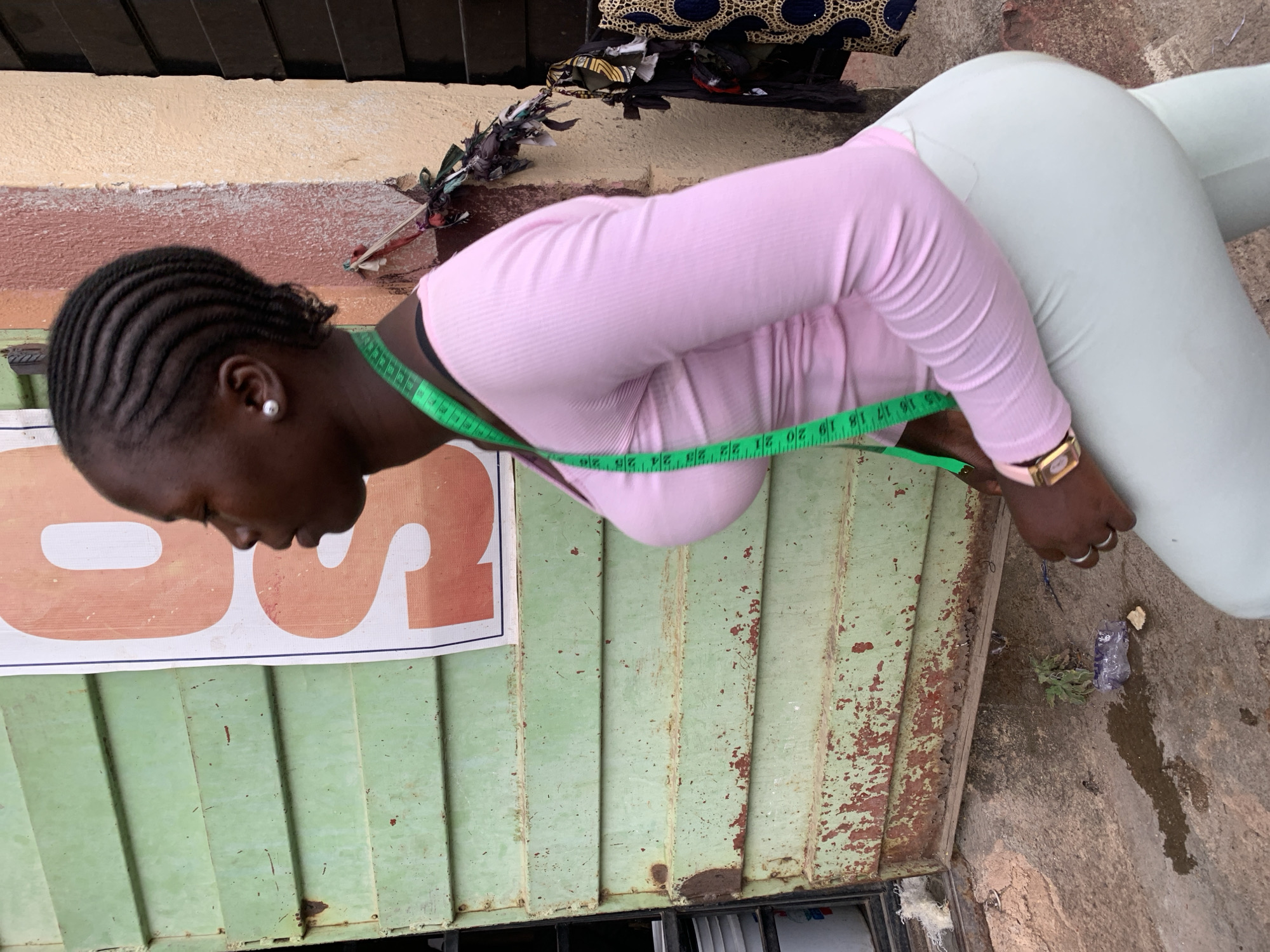 A medium shot shows a woman with her head bent down and a measuring tape around her neck. She is wearing a pink long-sleeved shirt and light green pants. She is standing next to a weathered green metal door with a white and orange sign attached to it. The background shows a beige wall and a stone surface. The location appears to be an urban area, possibly Jos, Nigeria.