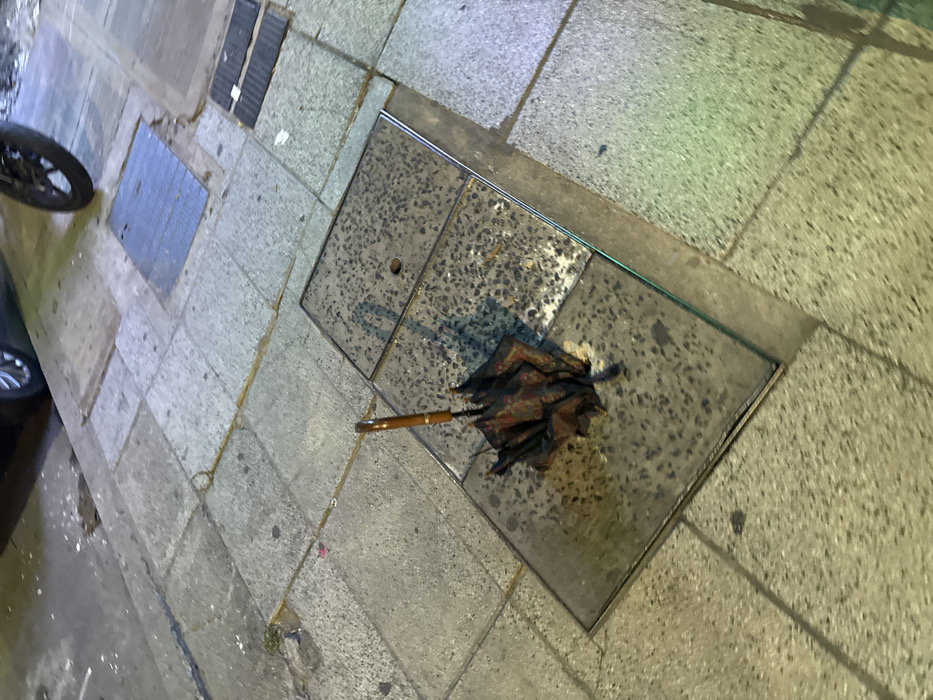 A discarded, damaged umbrella lies on a square concrete slab on a paved sidewalk. The umbrella, featuring a wooden handle, is dark and appears tattered, suggesting it has been through rough weather. The concrete slab is speckled and likely a utility access point, contrasting with the surrounding smooth pavement. A portion of a car tire is visible on the left side of the image. The overall scene suggests urban abandonment.
