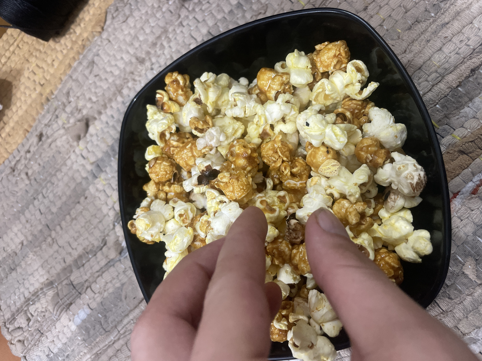 A close-up, overhead shot shows a black bowl filled with caramel and plain popcorn. A person's hand is reaching into the bowl, with fingers grasping several pieces of popcorn. The popcorn is arranged unevenly within the bowl, with some pieces appearing more caramelized than others. The bowl rests on a textured, woven surface, possibly a placemat or rug, which has a neutral, earthy tone. The background is slightly out of focus, but appears to be a dimly lit indoor setting. No specific landmarks or details indicating the location of Bhagsu, India are visible.