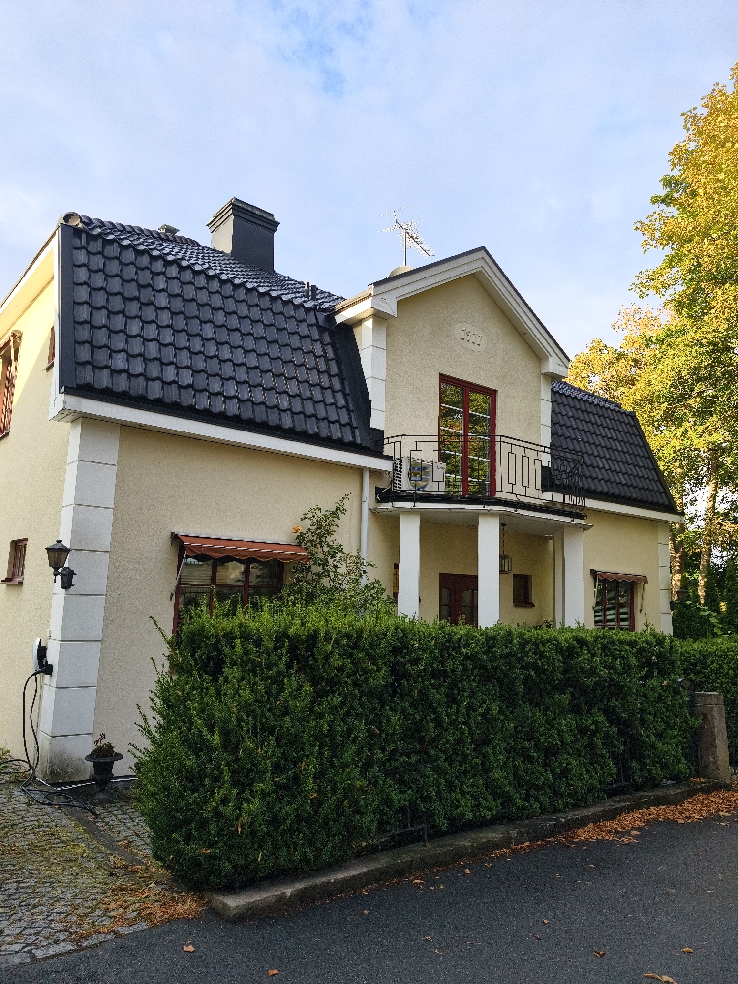 The image depicts a two-story, light yellow house with a dark gray tiled roof and a prominent black chimney. The house has a distinctive architectural feature: a small balcony with a black wrought-iron railing, supported by two white pillars. A year, "1911," is embossed in an oval shape above the balcony. Red-framed windows and a red-colored front door are visible. To the left of the house, a black outdoor lamp is mounted on a decorative pillar, and an electric car charging station is attached to the wall. A well-maintained, dense green hedge runs along the front of the property, separating it from a paved driveway and a street. Autumn leaves are scattered on the ground, suggesting the season. The sky is partly cloudy with patches of blue. The overall scene is a residential exterior in what appears to be a suburban or well-kept urban area. While the image is described as being in Norrköping, Sweden, no specific landmarks within the city are identifiable. There are no people present in the image.