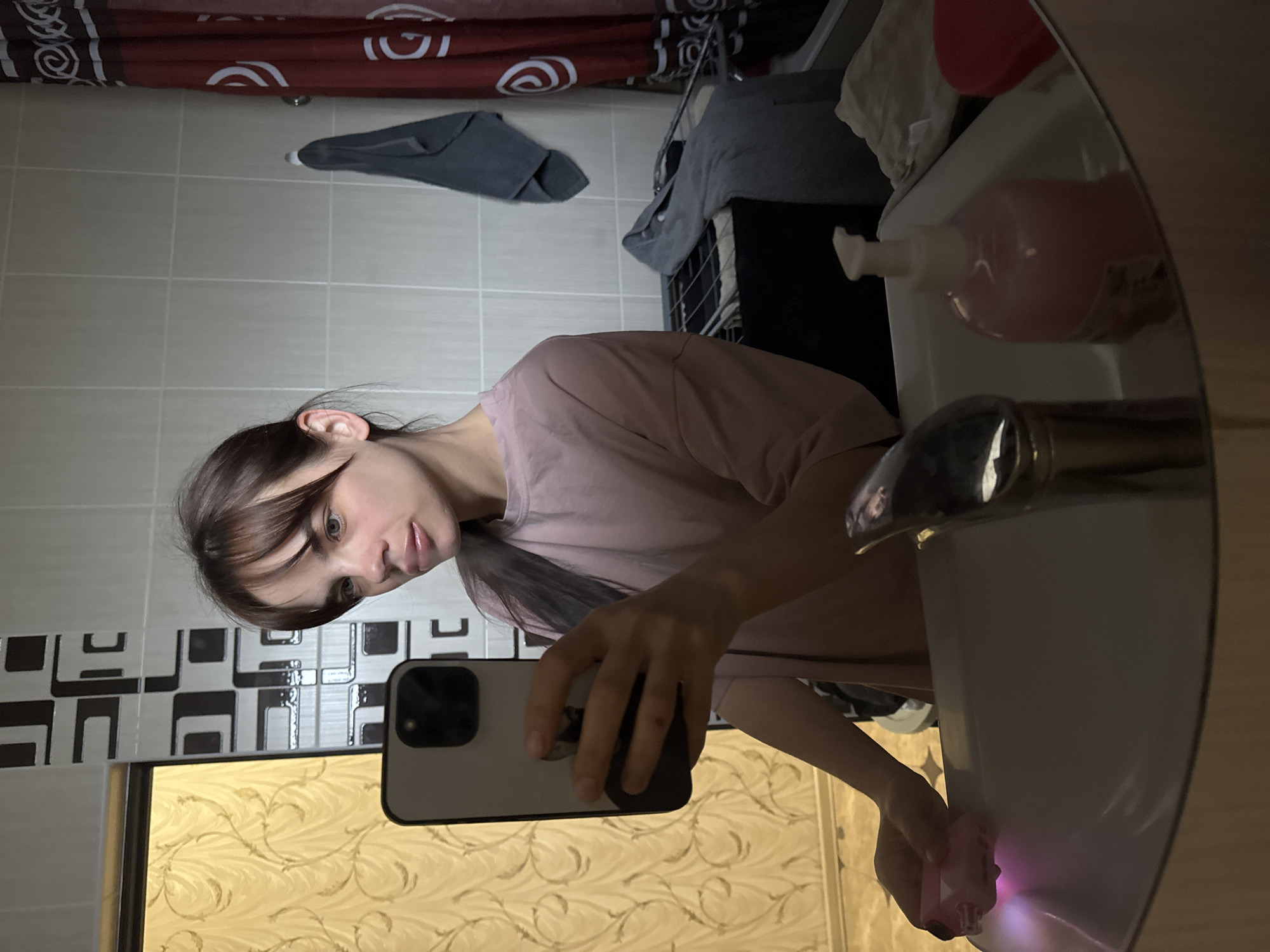A young woman is taking a selfie in a bathroom. She is facing a mirror, holding a smartphone in her right hand. The bathroom has patterned tiles on the walls and a sink with a pink soap dispenser. A towel rack with towels is visible in the background. The location is Rostov-on-Don, Russia.