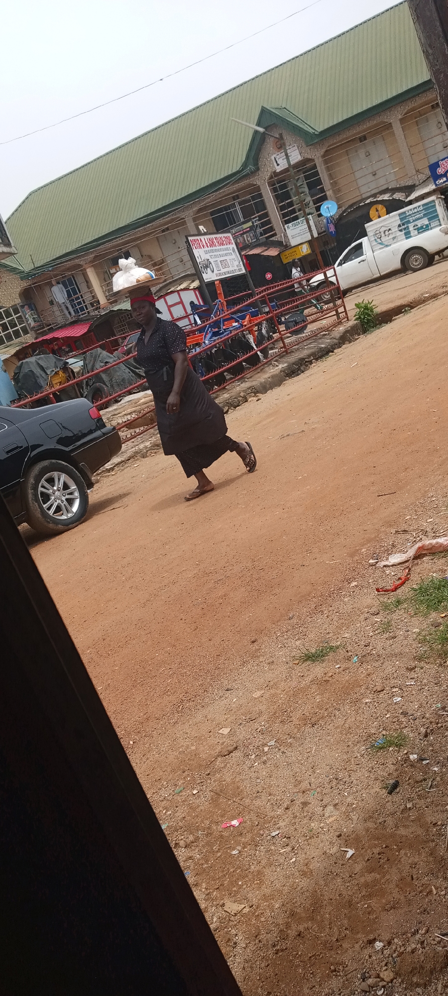 A woman is walking on a dirt road in what appears to be a commercial area. She is carrying a tray of items on her head. A black car is parked to her left. In the background, there's a building with a green roof and various shops and businesses. A pickup truck is also visible in the background. The scene is likely in a bustling part of the city of Jos, Nigeria, given the commercial activity and general environment.