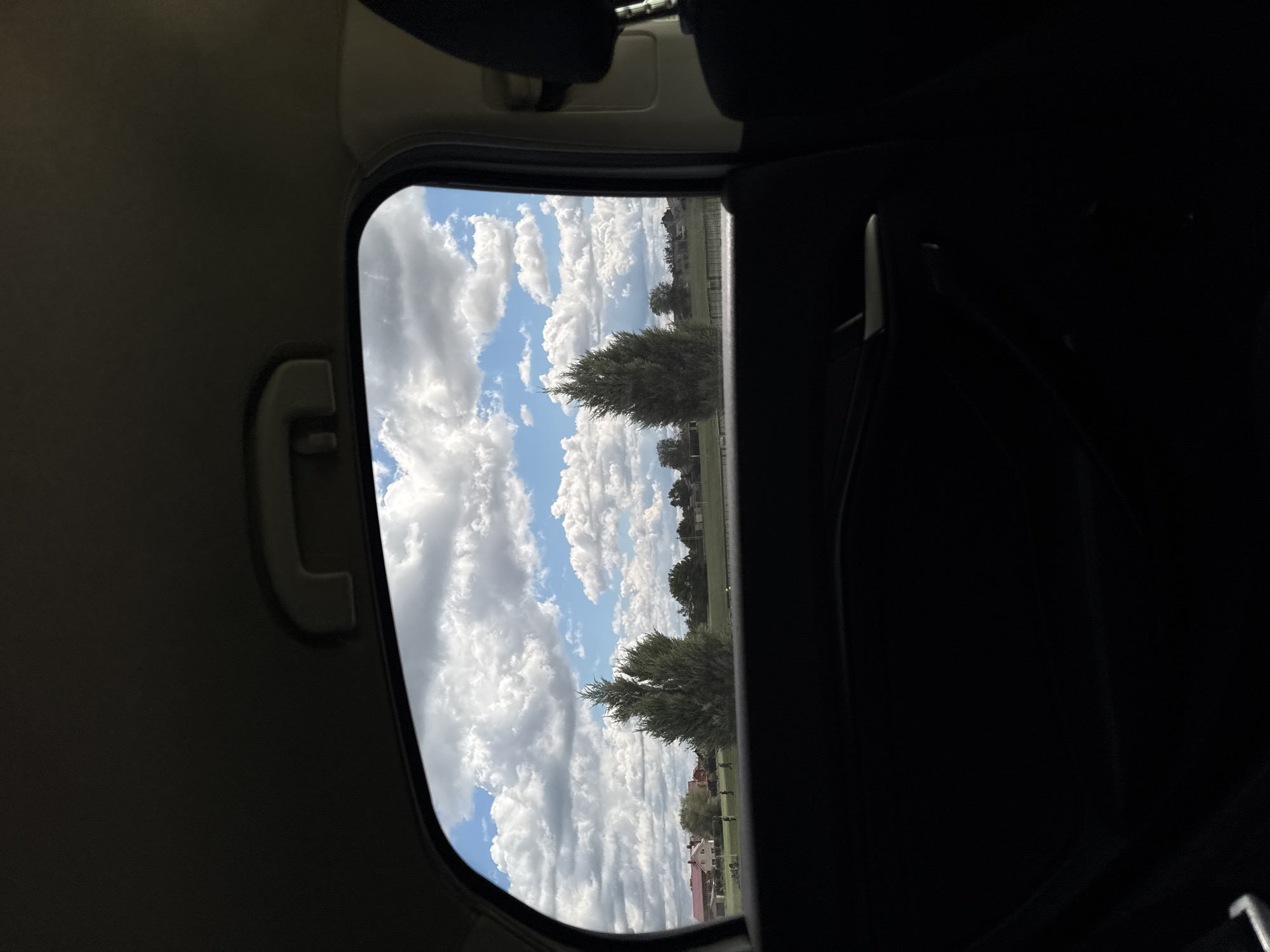 The view from inside a vehicle through a partially open window shows a daytime scene. Fluffy white clouds dot a blue sky. Trees, possibly evergreens, are visible in the middle ground. A building, possibly a house with a red roof, is seen further away. The interior of the vehicle, including a handle and a seat, is visible in the foreground. The location is Unknown, Unknown.