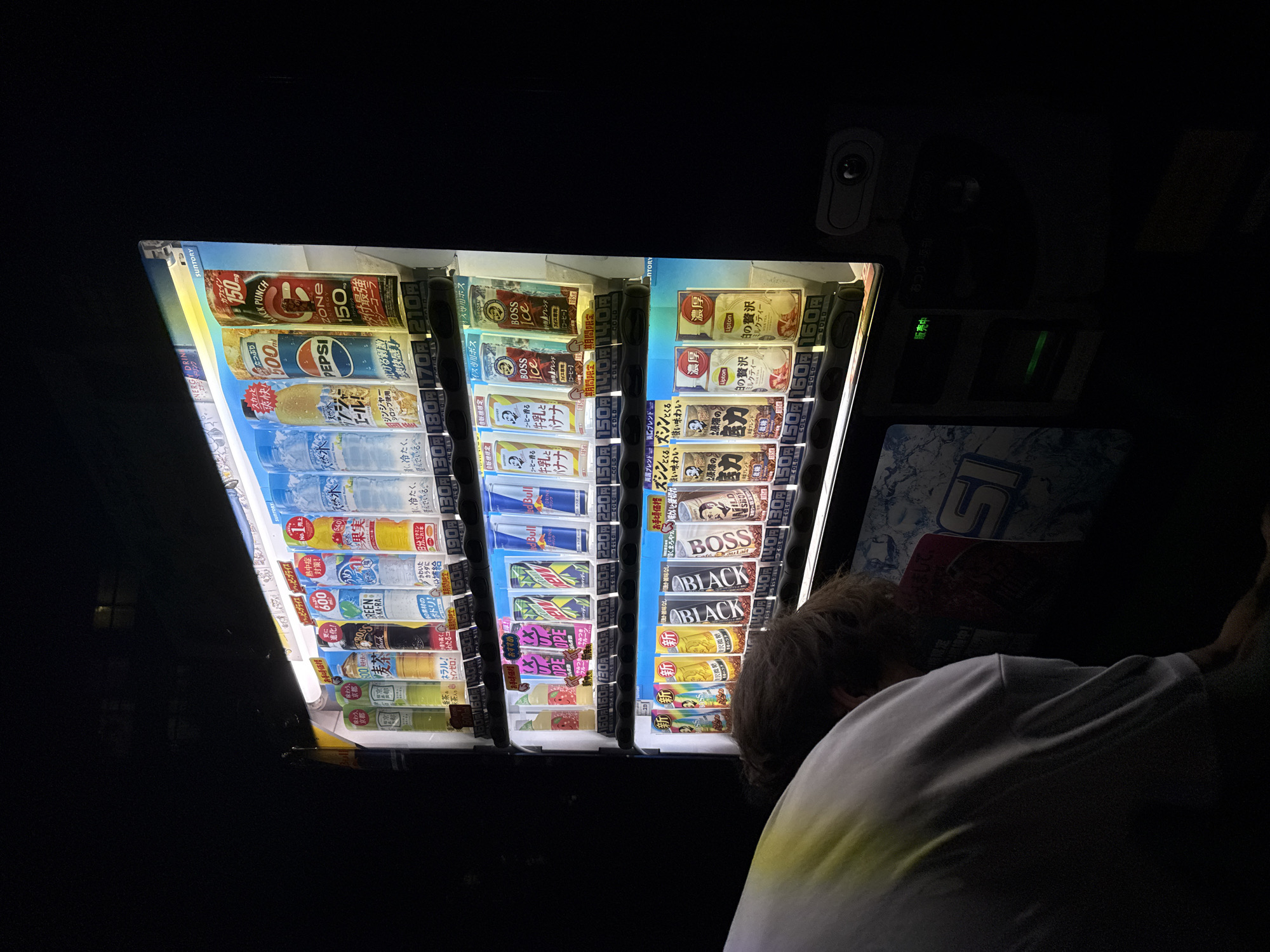 The image shows a person hunched over in front of a brightly lit vending machine in what appears to be a dimly lit area. The vending machine dispenses various beverages, including what seem to be bottled water, sodas, and canned drinks. Several brands are visible, such as Suntory, Pepsi, and Red Bull. The prices for the drinks are displayed below each item. The person's back is to the camera, and they are wearing a white t-shirt with a yellow and green gradient design on the shoulder. The vending machine has multiple tiers of products. The background is dark, suggesting it might be nighttime or an indoor, unlit space. The overall scene conveys a sense of everyday life, with the person interacting with the vending machine. The location is identified as Yachiyo, Japan.