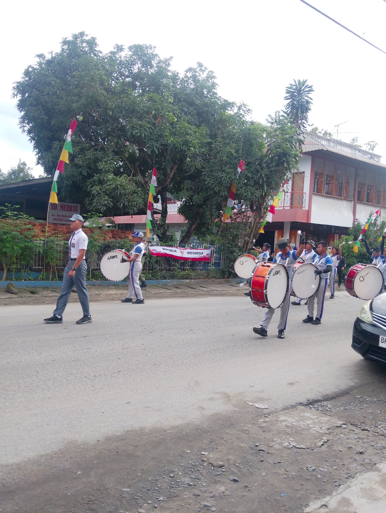 In the city of Serbelawan, Indonesia, a marching band composed of young individuals is parading down a paved road. The band members are dressed in white shirts, grey pants, and caps, with some carrying large drums. The parade is decorated with colorful flags, including the Indonesian flag, red and white. In the background, a building with a red roof and balconies is visible, surrounded by trees. The road is lined with more trees and a fence, suggesting a suburban or semi-rural setting. A black car is parked on the right side of the road, with its front bumper and headlights visible. The overall atmosphere appears to be festive, likely an Independence Day celebration or a similar public event.