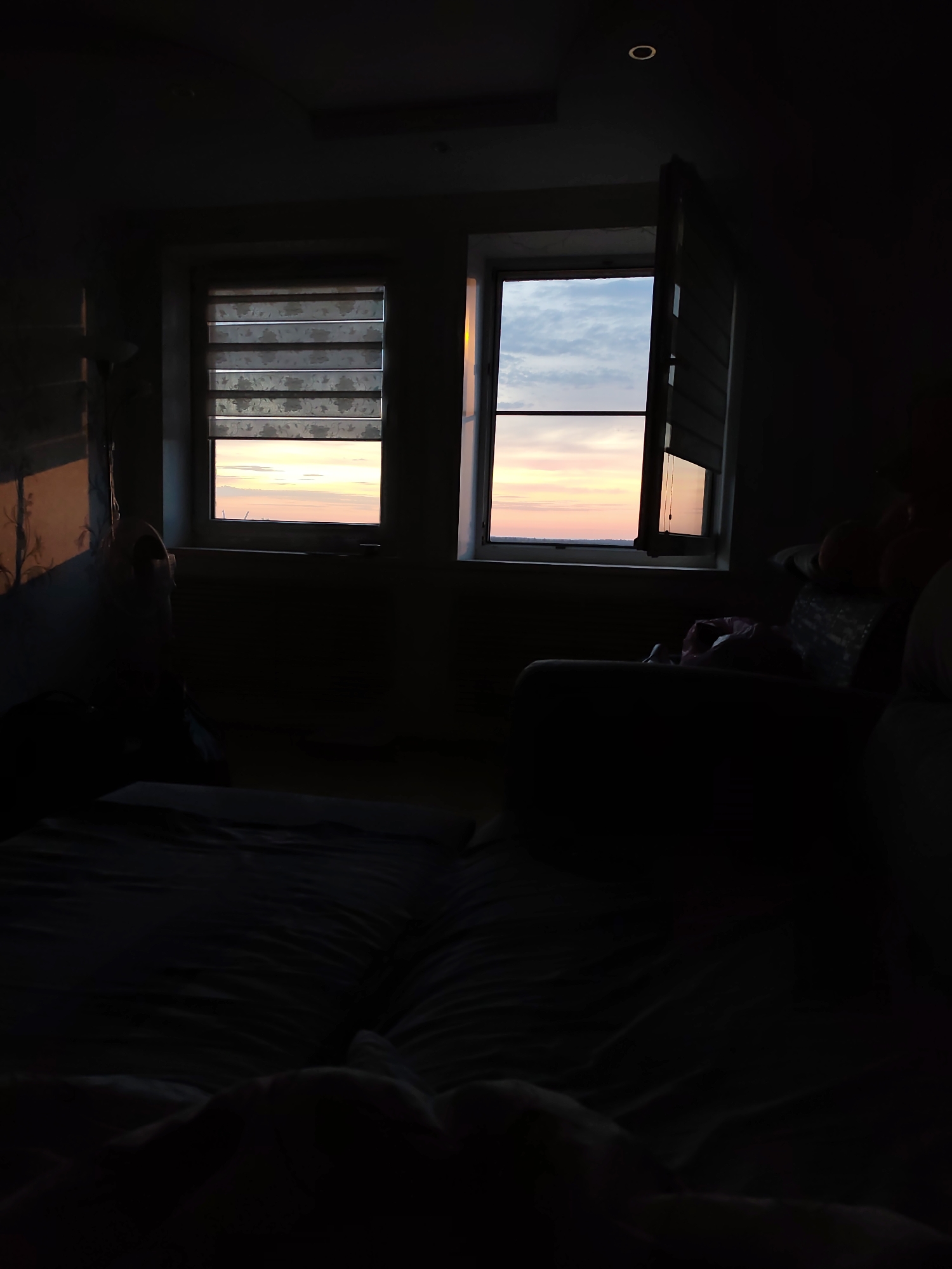 The interior of a darkened room is captured at dusk or dawn, as suggested by the colors visible through two windows. The window on the left has a partially drawn blind, revealing a sliver of the sky with hues of pink and orange. The window on the right is open, providing a clearer view of the sky's gradient of colors. A bed with rumpled covers occupies the foreground. To the right of the windows, an armchair or sofa is partially visible. The lighting is dim, with the natural light from the windows providing the main source of illumination. A single recessed light fixture can be seen on the ceiling. The image does not show any people. The location is not discernable as Gremyachinsk, Russia based on the image alone.