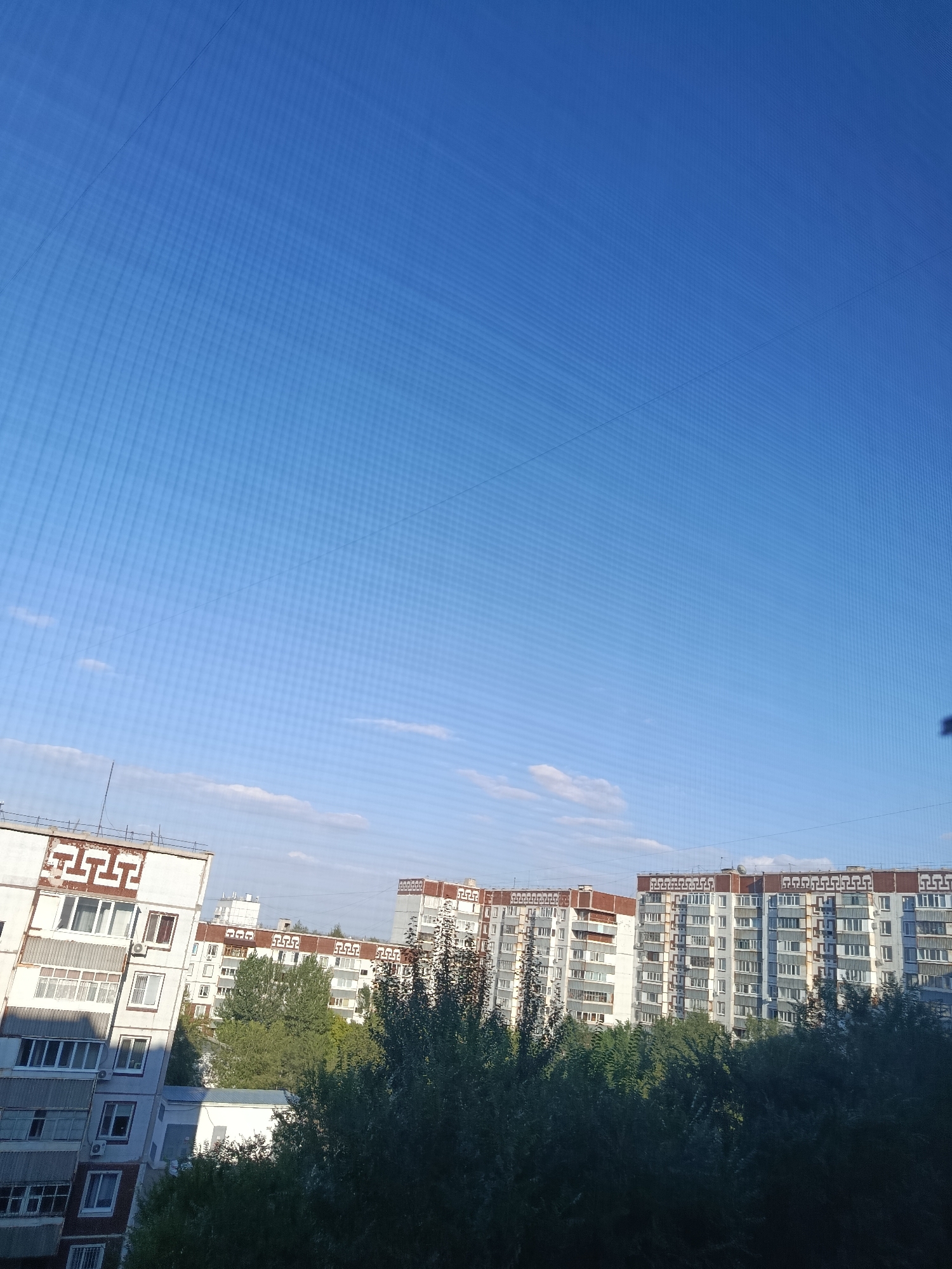 The image depicts a view of apartment buildings against a clear blue sky in Ekibastuz, Kazakhstan. The foreground is dominated by the upper floors of several residential buildings, characterized by their multi-story design with numerous windows and balconies. Some buildings feature decorative patterns along their facades. Lush green trees and foliage are visible in front of and between the buildings, suggesting a residential area with some greenery. The sky is mostly clear, with a few wispy clouds present. The overall impression is a typical urban residential landscape.