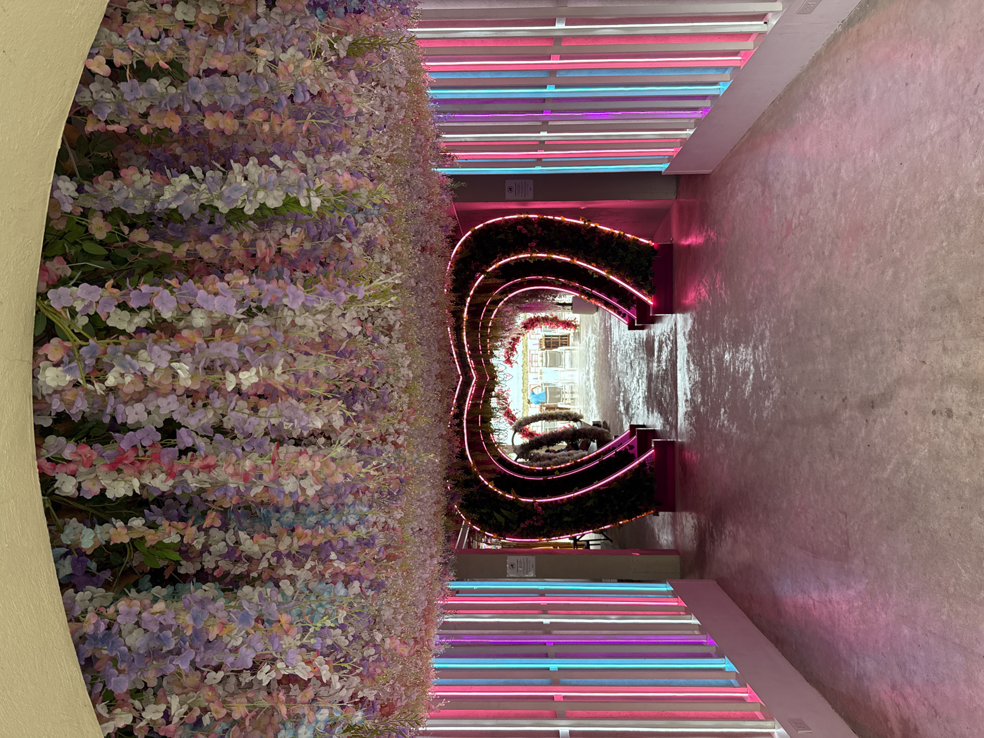 A dimly lit walkway is adorned with an overhead canopy of cascading artificial flowers in various pastel shades. On either side of the path, vertical rows of pink, blue, and white neon lights create a vibrant glow. In the foreground, the walkway opens up to a series of heart-shaped arches covered in greenery and outlined with pink neon lights, leading further into the distance. The ground is paved with a light-colored concrete. In the background, beyond the arches, a few tables and chairs are visible, with a glimpse of an outdoor area and a person wearing a blue shirt. The overall ambiance is romantic and somewhat whimsical.