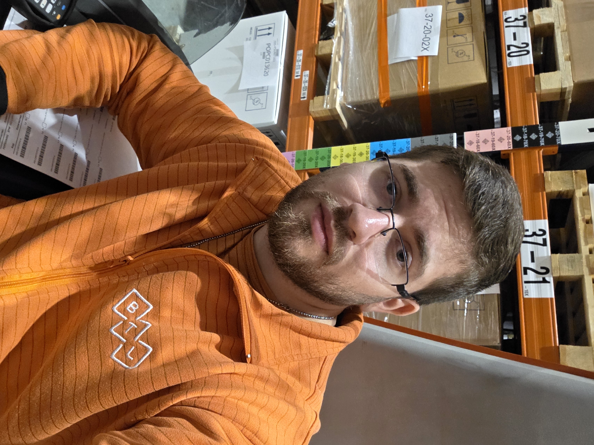 The photograph shows a man with a beard and glasses, wearing an orange striped zip-up hoodie, in an indoor environment. Behind him is a storage racking system containing boxes. Some of the labels on the racks and boxes are visible, with codes such as "37-21", "37-20", "37-20-02X", "POPC013620", and other alphanumeric strings. The man appears to be near a surface of some kind, possibly a desk or counter. There is no indication of the location being in Plovdiv, Bulgaria.