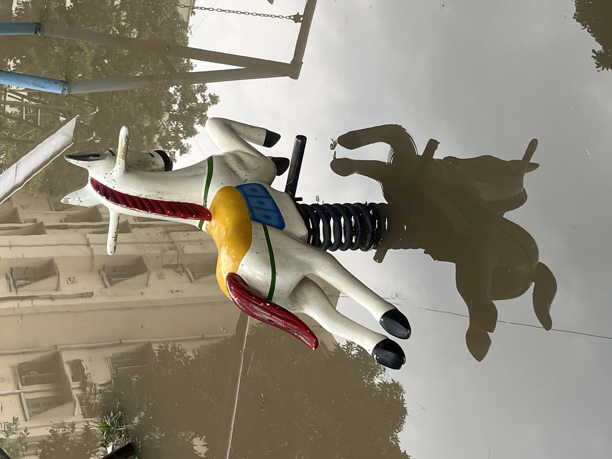 The image depicts a child's playground spring rider, shaped like a white horse with red, yellow, and blue accents, sitting partially submerged in floodwater. The horse's legs are black, and it is connected to a black spring. The dark reflection of the horse is visible in the water. Behind the horse, the image reflects buildings, trees, and a chain-link fence, suggesting the urban environment of Dhaka, Bangladesh.