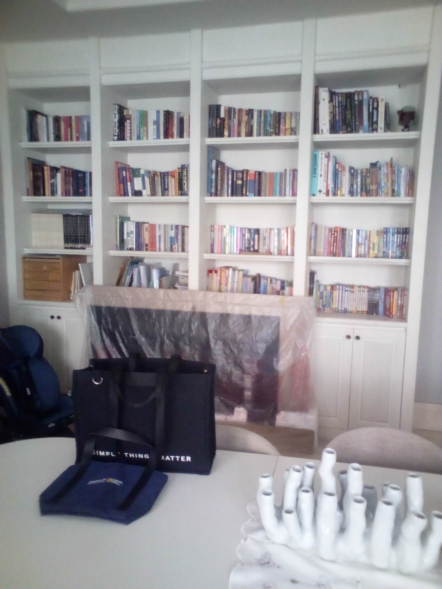 The image shows an indoor scene, likely a living room or study, featuring a large, white built-in bookshelf filled with numerous books and some decorative items. To the left of the bookshelf, a blue chair is partially visible. In front of the bookshelf, a large flat-screen television is covered in clear plastic sheeting. A small wooden chest of drawers is situated on the bottom shelf of the leftmost section of the bookshelf. On a white table in the foreground, there are two black tote bags and a blue cloth bag. A white decorative object, resembling coral or a cluster of tubes, is also on the table. The overall lighting suggests an indoor setting during the daytime. The image does not contain any elements that would allow for the specific location within South Jakarta, Indonesia, to be determined.