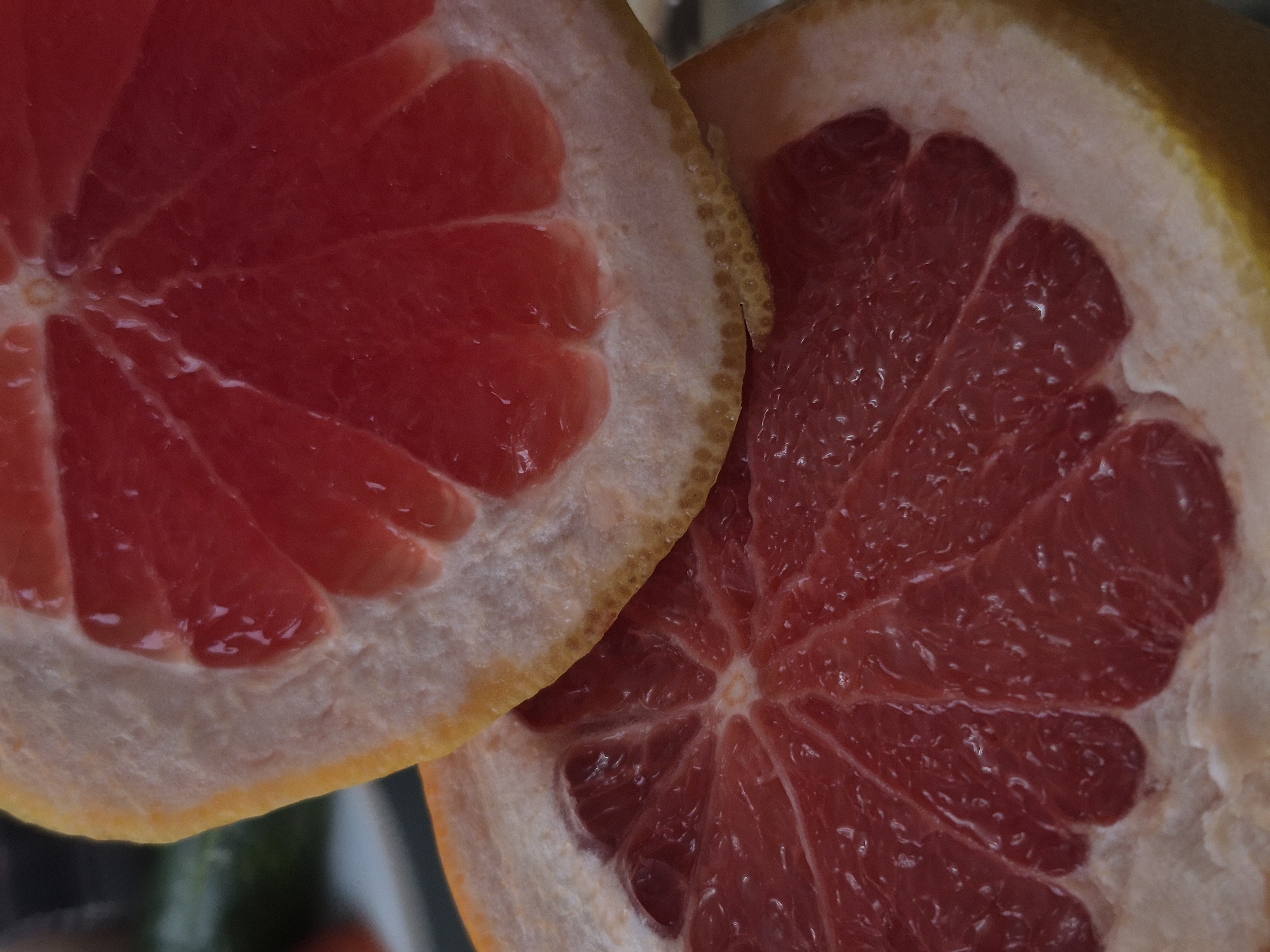 The media file presents a close-up, high-angle shot of two halves of a grapefruit, cut cross-sectionally. The dominant feature is the vibrant, ruby-red flesh of the fruit, which occupies most of the frame, displaying its distinct segments and juicy pulp. Individual juice vesicles and glistening moisture are visible on the surface of the fruit, suggesting freshness. Surrounding the red interior, the thick, pale white pith is clearly discernible, bordered by a narrow strip of textured yellow-orange rind.
The left grapefruit half is positioned slightly above and in front of the right half, creating a sense of depth and a dynamic composition. The background is softly blurred and indistinct, appearing dark with hints of green or other muted colors, ensuring the focus remains entirely on the intricate details and rich colors of the fruit. The lighting is even and soft, accentuating the natural textures and hues without harsh shadows. No activity is depicted; the image is a still-life composition emphasizing the aesthetic qualities of the grapefruit. There are no visible people, text, or specific location cues within the image itself. The scene appears to be an indoor setting due to the controlled lighting and lack of outdoor elements. The time of day and weather are not discernible.