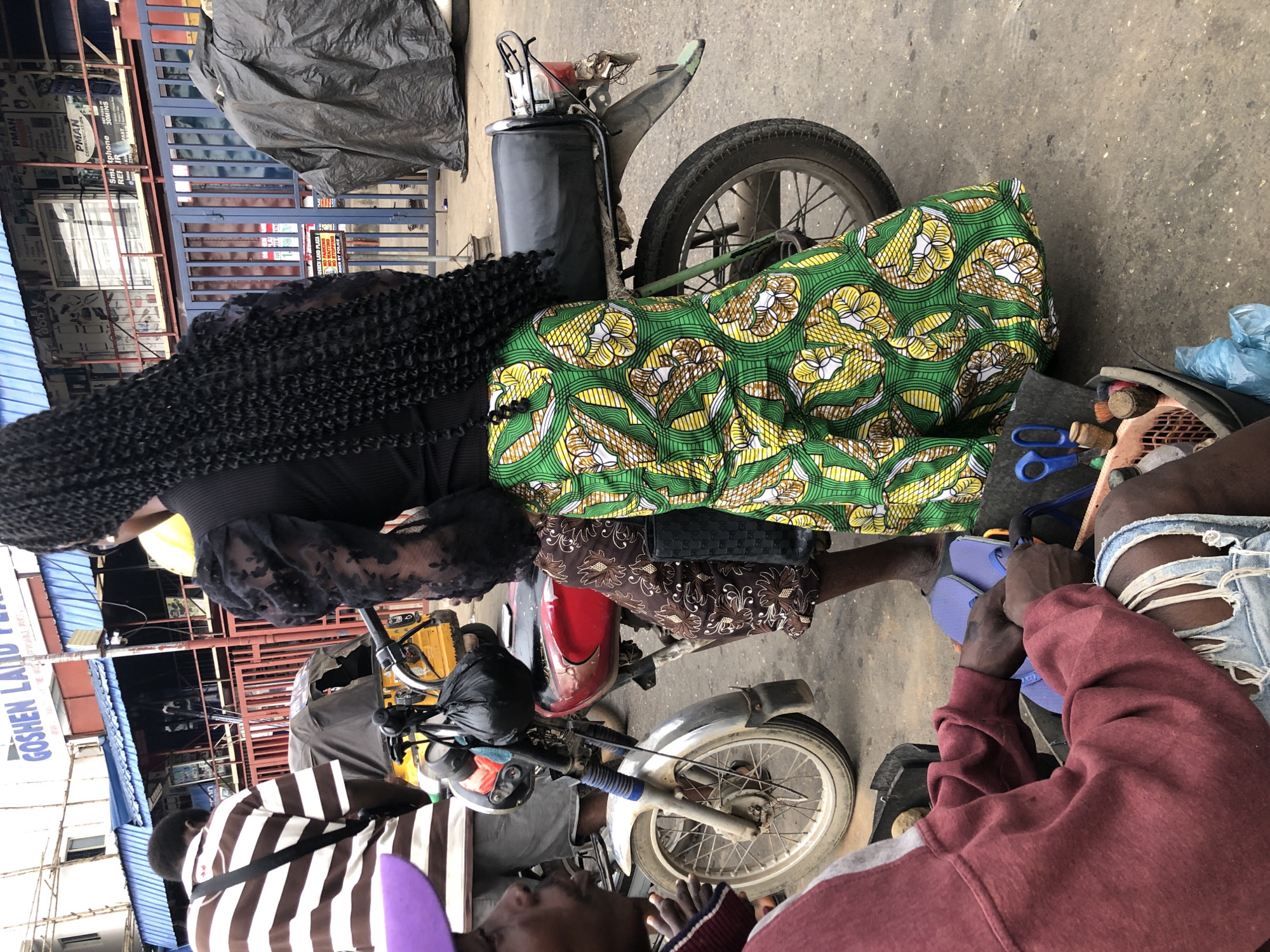 In Ikeja, Nigeria, a street scene unfolds around a parked motorcycle. A person with long, dark braided hair, wearing a black top with puffed sleeves and a vibrant green and yellow patterned skirt, stands near the motorcycle. To the left, another individual, wearing a maroon hoodie and distressed denim shorts, is seated, seemingly engaged in a task. Their hands are visible, manipulating items within a basket containing tools and blue flip-flops. In the background, a yellow and red motorcycle is partially visible, with a black bag hanging from its handlebars. Buildings with signage, including "GOSHEN LAND PLAZA," are present in the urban environment, suggesting a commercial area.