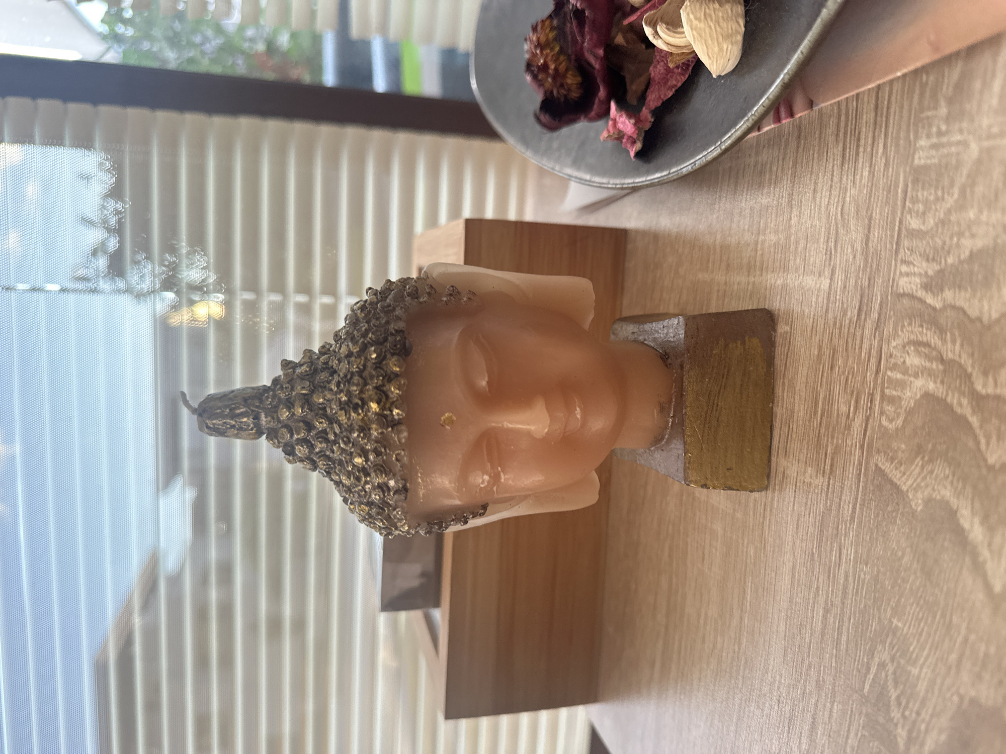 A close-up, eye-level shot captures a Buddha head candle on a light brown wooden surface. The candle is a pale peach color, with a detailed, gold-colored headdress that resembles intricate curls. A small, gold-colored mark is visible on the forehead, between the eyes. The base of the candle is a rectangular block, painted gold. Behind the candle, a light brown wooden box with a slightly darker rectangular insert sits on the wooden surface. To the right of the candle, a dark gray plate holds dried, reddish-brown flower petals and light brown, almond-shaped objects. The background features a window with horizontal blinds, allowing diffused light to enter. Reflections of outdoor foliage and a bright sky are visible on the blinds. The scene is indoors, likely in a home or commercial establishment in Sozopol, Bulgaria.