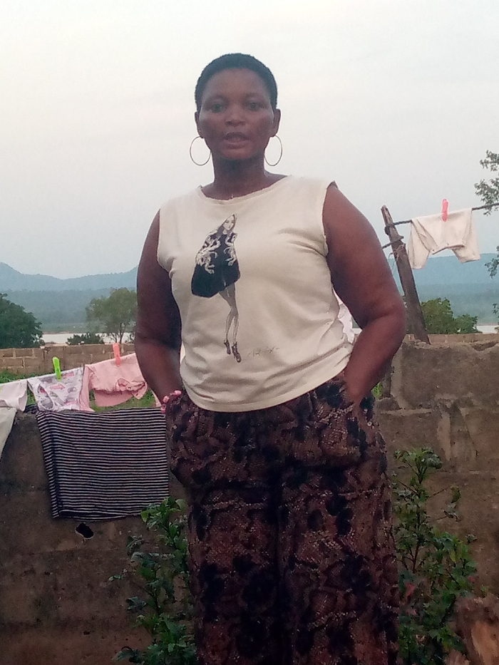 A medium shot shows a dark-skinned woman with short hair and hoop earrings standing outdoors. She is wearing a sleeveless cream-colored t-shirt with a graphic of a woman in a black dress and patterned, dark-colored, loose-fitting pants. Her right hand is in her pocket, and she is wearing a pink beaded bracelet on her left wrist. Behind her, a clothesline is strung with laundry, including white undergarments clipped to a wooden pole with a pink clothespin. To the left of the clothesline, several pieces of clothing, including a pink garment and a striped garment, are draped over a wall. In the background, rolling hills and trees are visible under an overcast sky. The scene appears to be in a rural or semi-rural area, possibly on the outskirts of Ajaokuta, Nigeria, given the natural landscape and the presence of drying laundry. The woman is facing the camera with a neutral expression. The overall lighting suggests it is either early morning or late afternoon.