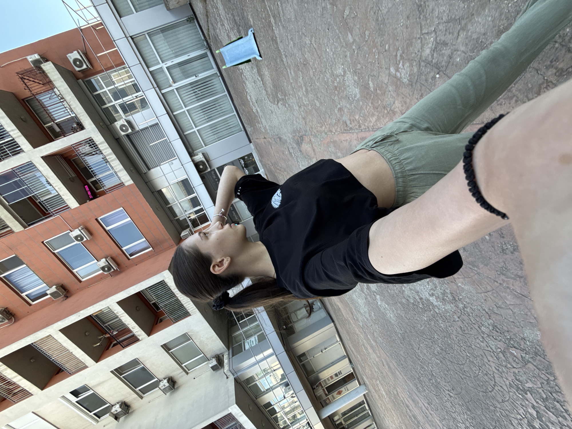 A young woman wearing a black T-shirt and green trousers is lying on a rooftop in Sofia, Bulgaria. She has dark hair pulled back in a ponytail. Her face is turned toward the sky, and her hand is raised to her forehead. Buildings are visible in the background, including a taller building with numerous windows and air conditioning units. A small blue object sits on the rooftop, which is a textured gray surface. The lighting indicates it is daytime.