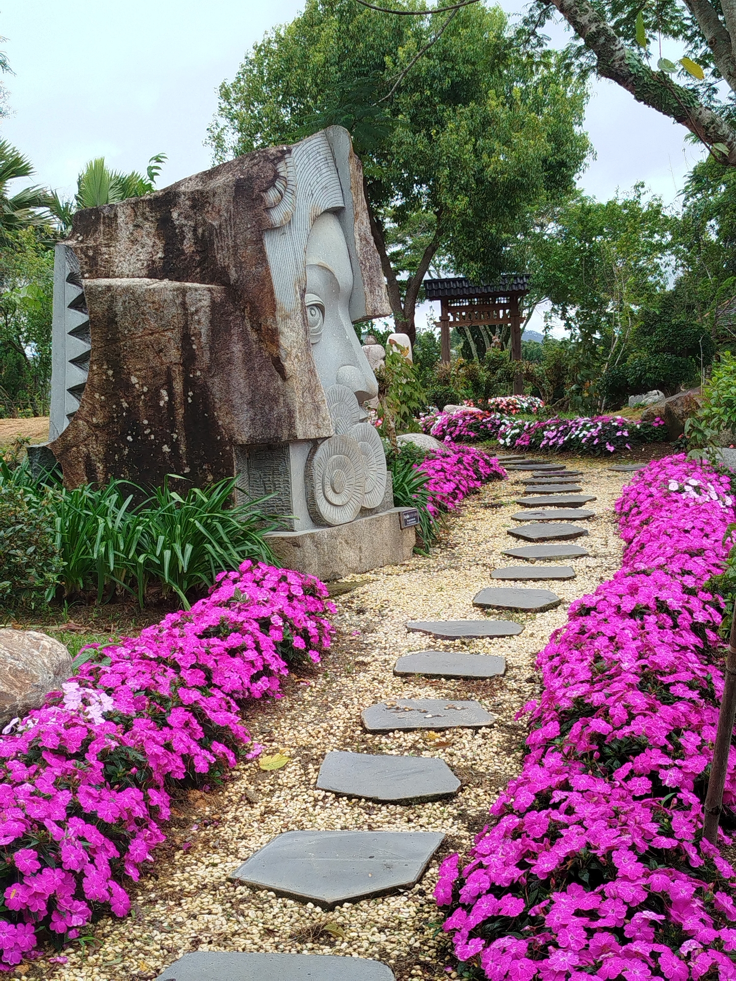 In Lâm Viên - Đà Lạt, Vietnam, a stone sculpture resembling a face is positioned in a garden. The sculpture is set against a backdrop of lush green trees and a cloudy sky. A pathway made of flat stones meanders through a vibrant bed of bright pink flowers, leading the eye towards the sculpture. The pathway is bordered on both sides by more pink flowers, creating a striking contrast with the earthy tones of the path and the stone sculpture. In the background, a traditional wooden structure with a tiered roof is visible amongst the foliage, adding to the serene and artistic atmosphere of the location.