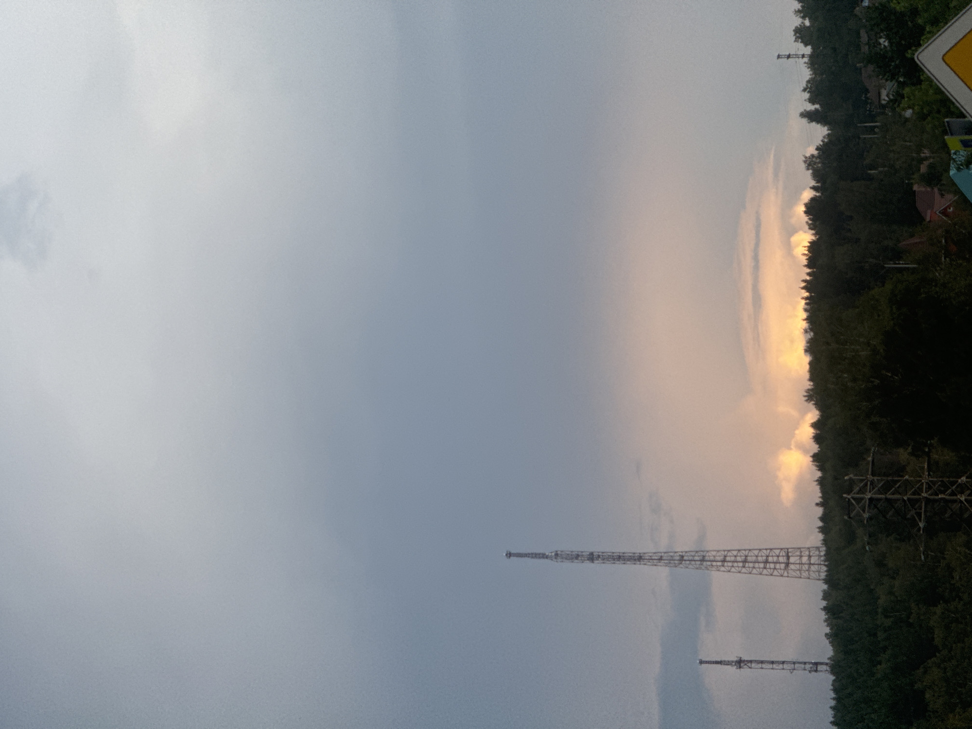 The image shows a panoramic view of the landscape with two prominent radio towers situated on a tree-covered hill. The sky is overcast with a soft, diffused light, suggesting either early morning or late evening. The radio towers, one taller than the other, stand stark against the cloudy sky, their metallic structures clearly visible. In the foreground, a dense forest covers the rolling hills, with the tops of trees forming a dark green line across the landscape. Some residential buildings and a power line are also visible amidst the trees, hinting at human habitation within this natural setting. The scene appears peaceful and quiet, with no people or animals present. The overall impression is one of a serene, rural environment, possibly on the outskirts of Markova, Russia, given the context.