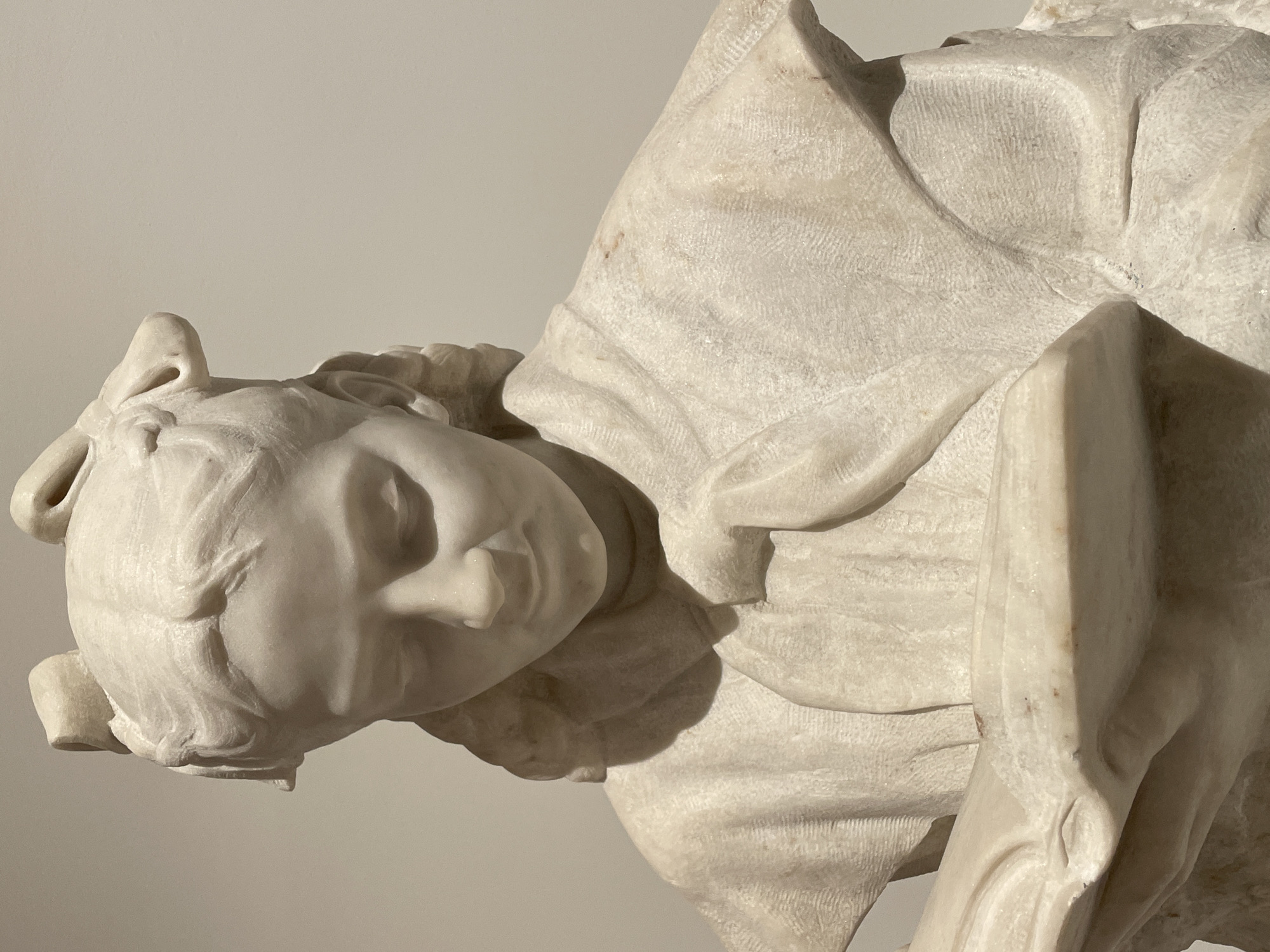 The image presents a finely detailed white marble bust of a young girl or woman engaged in reading. The figure is depicted from the upper chest, with her head gently bowed, eyes cast downwards, focused intently on an open book held in her hands. Her hair is intricately sculpted, featuring two distinct bows or ribbons positioned on top of her head, with braided or styled hair flowing down the sides. She wears a garment resembling a cape or shawl, characterized by rendered fabric folds and textured surfaces. The white marble material shows subtle natural veining and isolated reddish-brown discoloration on the right shoulder and near the book. The sculpture is illuminated from the top-left, creating soft shadows under the chin, nose, and along the garment's folds. The background is a plain, light beige wall, indicative of an indoor gallery or museum setting. This artwork, titled "The Reader" by Vasily Kononov, is located at the State Tretyakov Gallery, Moscow, Russia.