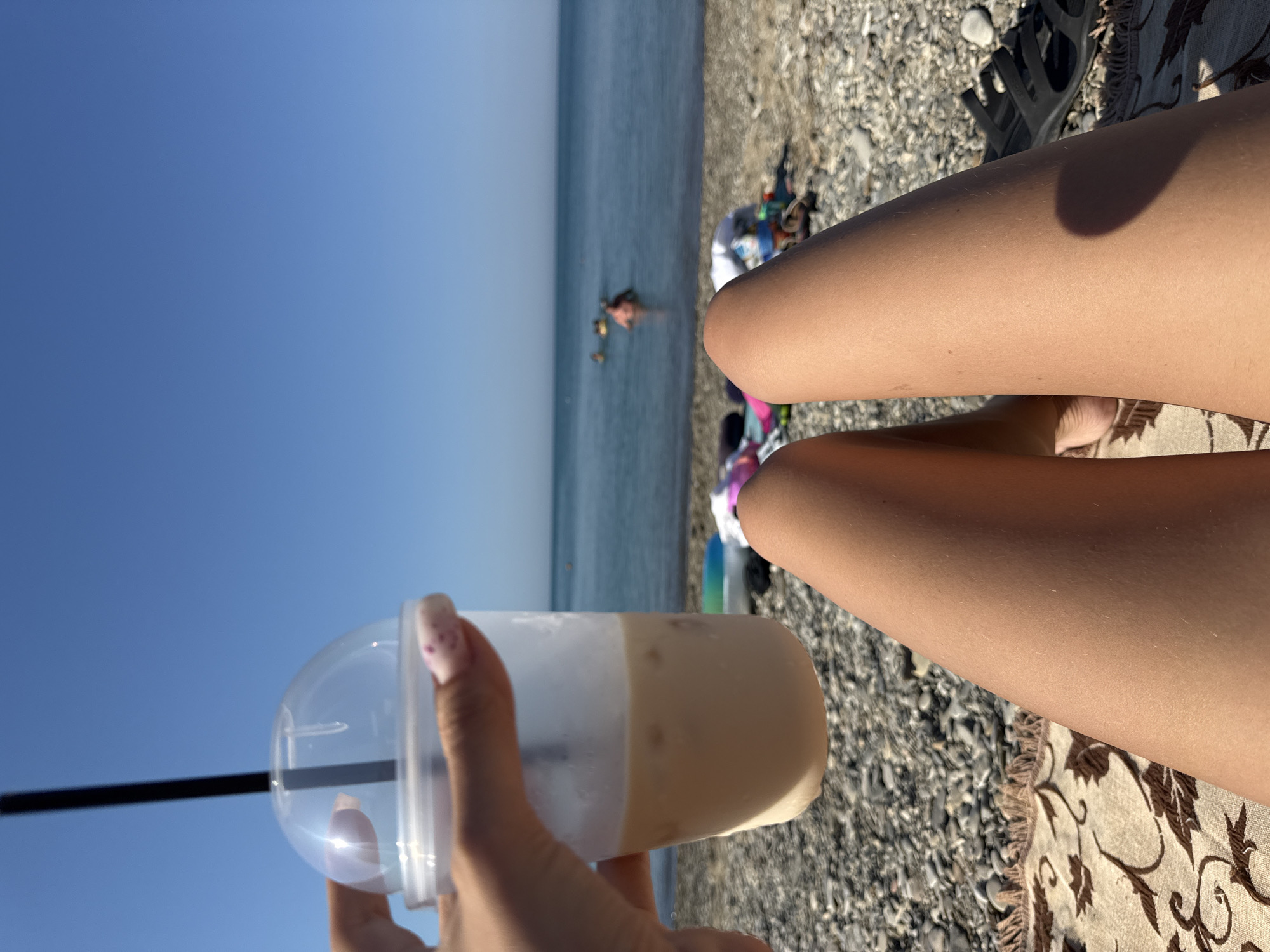 The image shows a close-up view of a person's legs and a drink on a pebble beach, with the sea and sky in the background. The person is lying on a patterned blanket, and their legs are tanned. They are holding a clear plastic cup filled with a light-colored beverage, possibly an iced coffee or latte, with ice cubes visible. A black straw extends from the cup. In the distance, several people are visible in the calm, blue sea. The sky is clear and bright blue. The beach is covered in smooth, grey pebbles. The overall scene depicts a relaxing day at the beach. The location is specified as the city of Dzhubga, Russia.