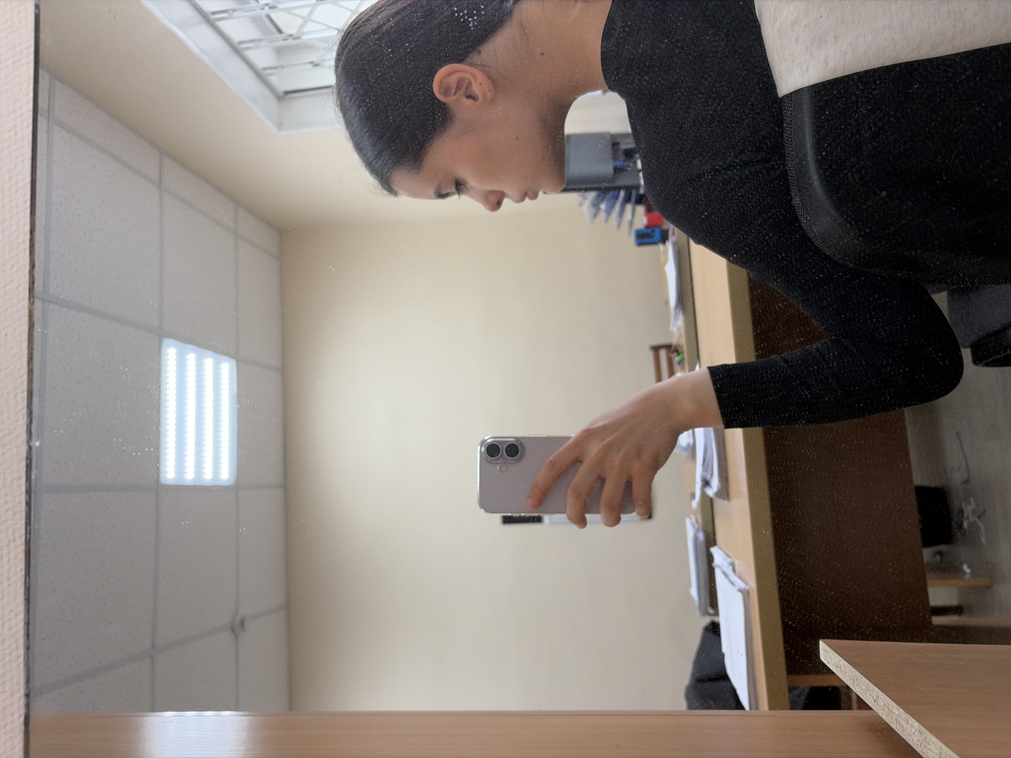 A person is taking a selfie in a mirror. The reflection shows a room with drop ceiling tiles and a fluorescent light fixture. A desk with papers and objects on it is visible to the right of the mirror. The location is likely in an office building or similar institutional setting in Chaikovsky, Russia.