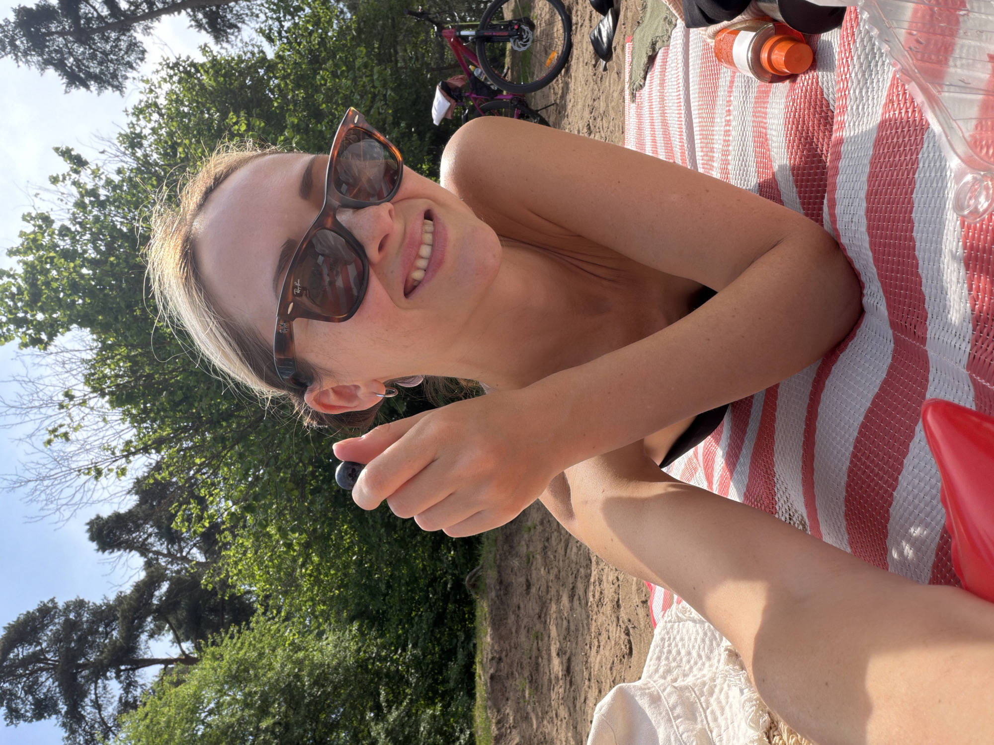 A woman is lying on a red and white striped beach mat outdoors. She is wearing sunglasses and holding a dark berry in her fingers, smiling slightly. Behind her are trees and a glimpse of a bicycle. The ground appears to be sandy. The scene suggests a casual, sunny day, likely at a park or beach. There is no information to determine if the location is the city of Moscow, Russia.
