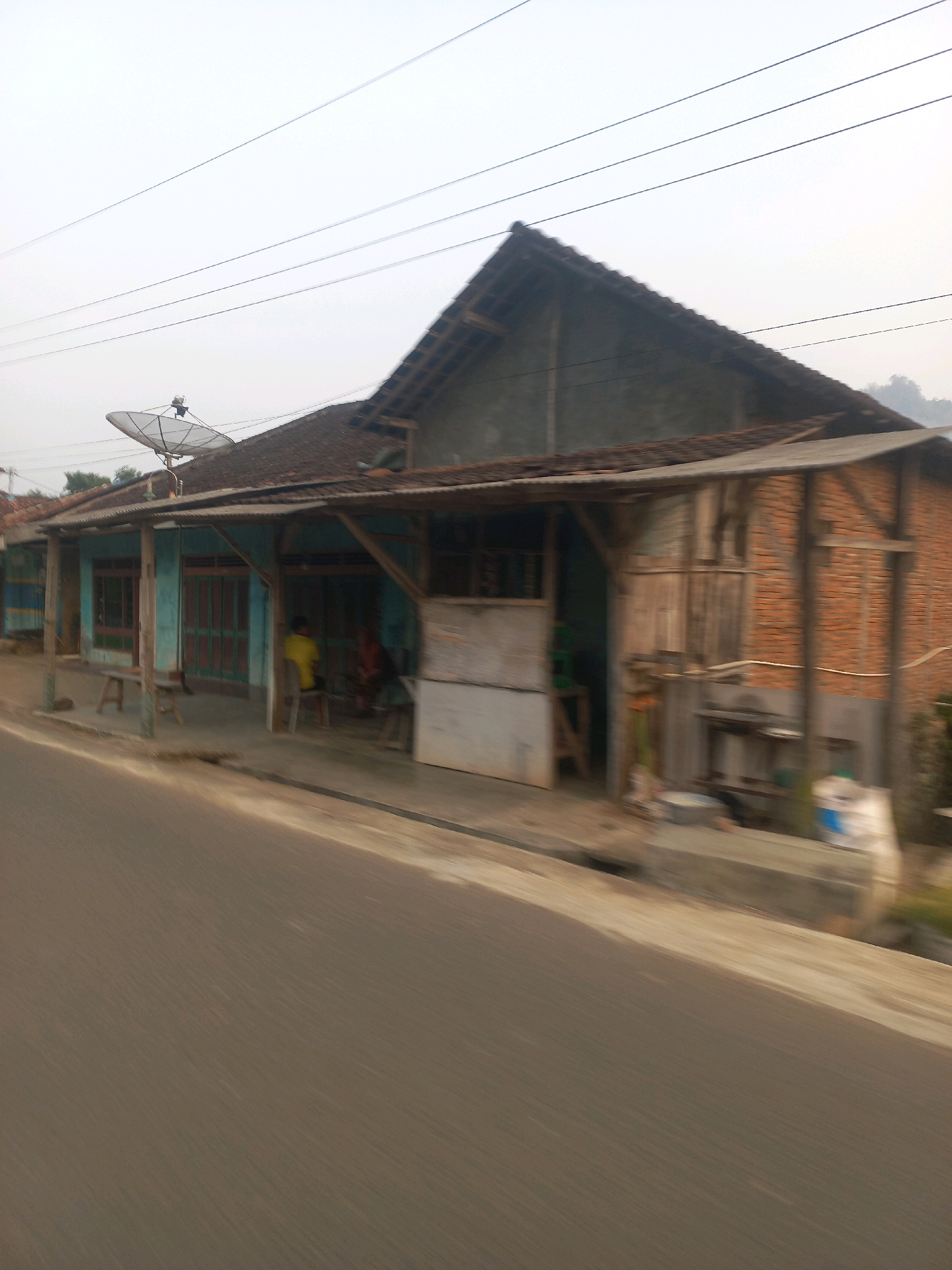 A low-angle, motion-blurred medium shot captures a roadside scene in Sukoharjo, Indonesia, under an overcast sky. The image is dominated by a weathered, open-fronted building with a sloping, dark tiled roof supported by rough wooden posts. The building's facade features a light blue painted wall with darker blue and green striped sections on the far left, and what appear to be red-framed glass doors or windows. A satellite dish is mounted on the roof.
To the right, a section of the building is constructed with visible reddish-brown brickwork. A rough wooden exterior wall is situated between the brick and the blue facade. Shelving and various objects, including buckets and bags, are visible within and around the building.
In front of the building, a raised concrete walkway or sidewalk runs parallel to a blurred asphalt road in the foreground. Several simple wooden benches or stools are placed along the walkway. Two figures are partially visible within the building's open front. One individual, wearing a yellow shirt, is seated on a stool. The other figure is seated further back and is less distinct. Electrical wires stretch across the upper portion of the frame against the pale sky. The overall impression is of a humble, perhaps commercial or residential, structure in a rural or semi-rural setting.