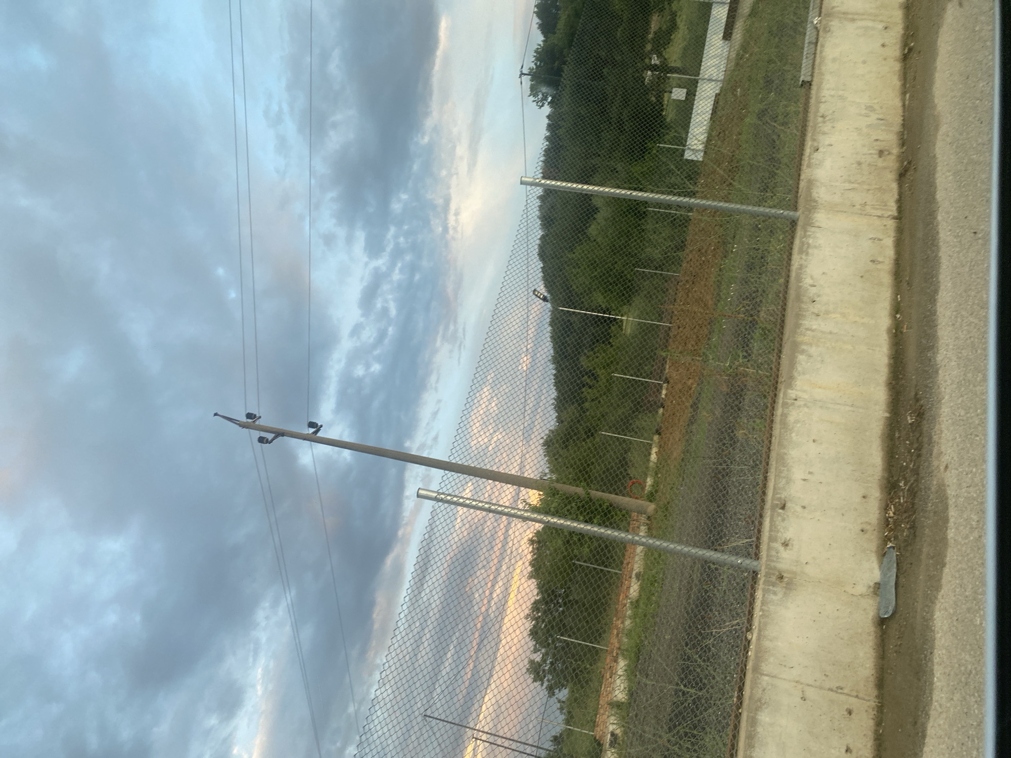The image depicts a rural or semi-rural scene with a prominent chain-link fence in the foreground, running horizontally across the frame. Behind the fence, there is a patch of reddish-brown earth, followed by a dense line of green trees forming a backdrop. The sky above is filled with dramatic, cloudy formations, suggesting either twilight or an impending storm. A single wooden utility pole with electrical wires extending from it is visible, positioned slightly off-center to the right. The lower portion of the image shows a concrete barrier, with some debris and a small, dark object on its surface. This scene is located in Ablanitsa, Bulgaria.