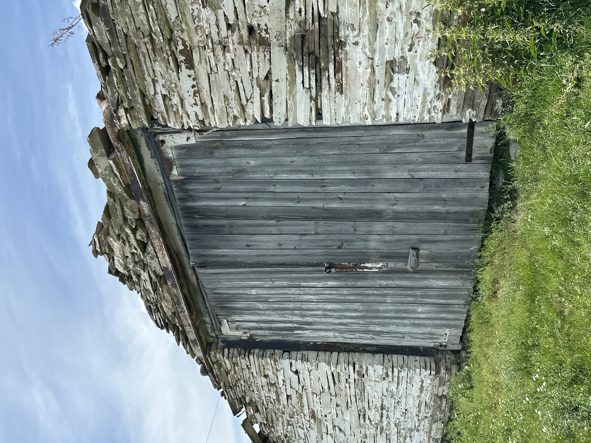 An old, weathered wooden barn door is set into a crumbling stone wall under a cloudy sky. The stone wall is constructed of irregular, flat layers of light-colored rock, with some sections showing peeling white paint and exposed mortar. The top of the wall is uneven, with loose stones and a rusty metal beam visible along the edge. The wooden door is made of vertical planks, grayed and aged, with a simple latch and a small wooden bar. A rusty hinge is attached to the right side of the door. The ground in front of the barn is covered with green grass and some small wildflowers.