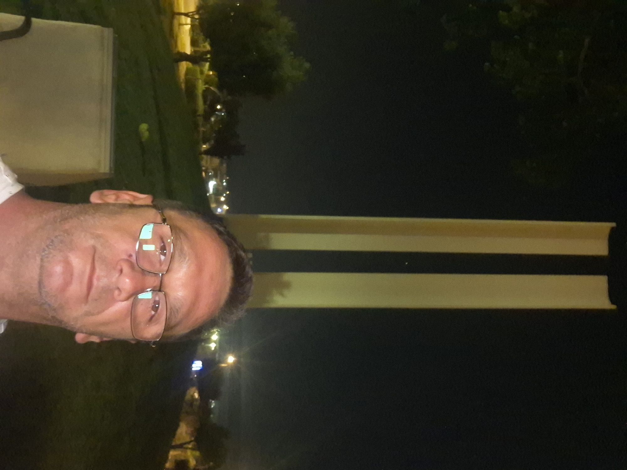 A medium shot photograph shows a man with short, graying hair and glasses standing in front of a night scene in Yerocham, Israel. The man is positioned in the lower half of the frame, looking directly at the camera with a neutral expression. He is wearing glasses with rectangular lenses and a light-colored shirt. In the background, two tall, slender, beige pillars stand vertically against a dark night sky. The pillars appear to be a prominent landmark, possibly a monument or architectural feature. To the left of the pillars, a park-like setting is visible, with trees and illuminated city lights in the distance. The ground in the park is covered with grass. The overall scene is dimly lit, characteristic of an outdoor night environment.