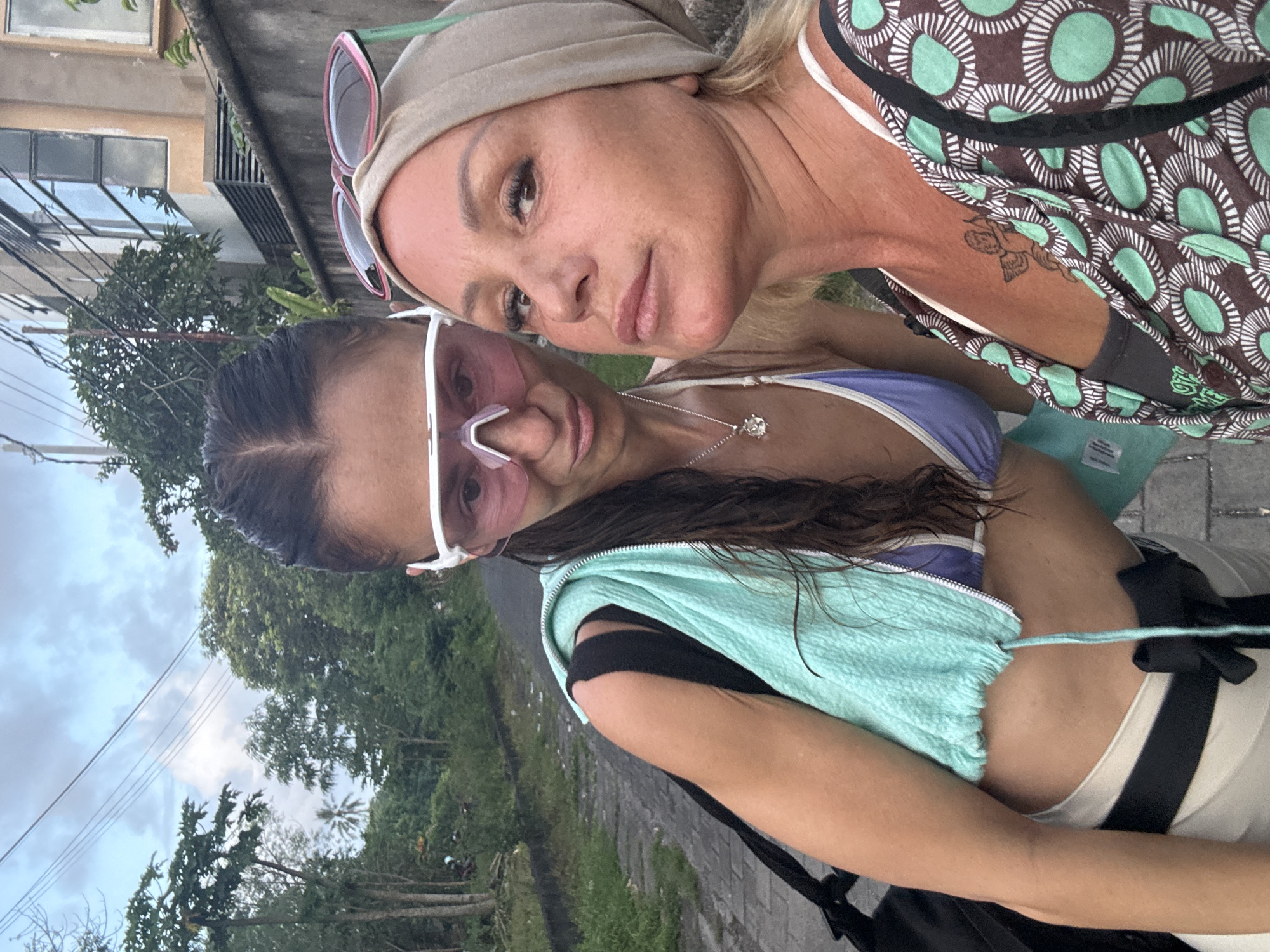 The original image is a close-up, selfie-style photograph featuring two women outdoors, rotated 90 degrees clockwise. The setting is likely Kuta, Indonesia, as indicated by the provided context.
In the foreground, a woman with dark hair wears distinctive pink-tinted sunglasses with thick white frames. She has on a light purple top, partially covered by a light blue textured garment, possibly a zipped vest or jacket, draped over her arm. A silver necklace with a circular, ornate pendant is visible around her neck. Her expression is neutral as she looks towards the camera.
Behind her, a second woman, with blonde hair, wears a light beige head covering and has pink and green sunglasses pushed up onto her head. She is dressed in a brown top featuring a bold pattern of concentric teal and white circles. A small, dark-inked tattoo is visible on her upper arm. She also looks towards the camera with a relaxed, slightly smiling expression.
The background reveals a tropical environment with abundant green foliage and trees. Utility lines crisscross against an overcast, cloudy sky, suggesting either a cloudy day or a time of day like early morning or late afternoon. Portions of buildings with windows are visible through the trees, hinting at an urban or semi-urban area. The ground appears to be a paved path or road. The overall mood is casual and friendly. No discernible text is clearly legible on any objects within the frame.
