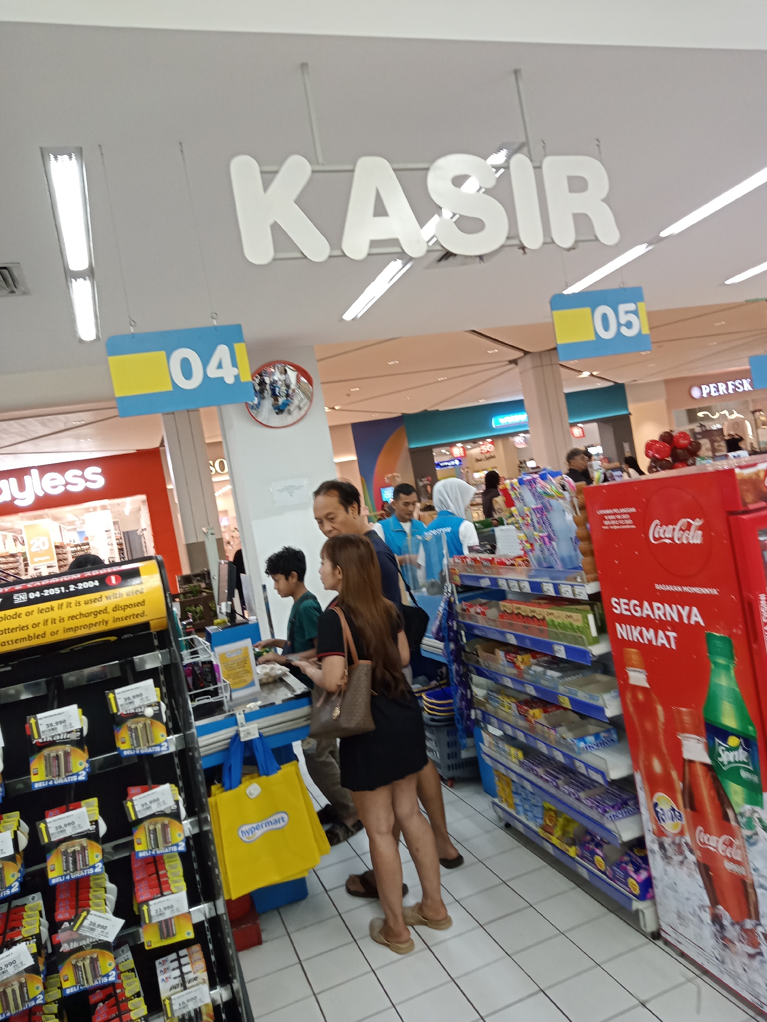 In a brightly lit, indoor retail environment, possibly a supermarket or department store, a checkout area is visible. Above the checkout lanes, large white block letters spell out "KASIR" (Indonesian for "Cashier"). To the left and right of the "KASIR" sign, blue and yellow signs display numbers "04" and "05" respectively, likely indicating lane numbers.
In the foreground, to the left, a display rack holds various battery packs. Further back, a woman wearing a black dress and carrying a brown purse stands at a checkout counter, interacting with a cashier. To her left, a young boy in a green shirt stands near the counter. A man, likely the woman's companion, stands behind her, looking towards the checkout.
To the right of the checkout area, shelves are stocked with various products, including drinks. A prominent red display board features the Coca-Cola logo and the text "SEGARNYA NIKMAT" (Indonesian for "Refreshing Enjoyment"). Other drinks like Fanta and Sprite are also visible. The floor is tiled in a white grid pattern. The overall impression is a typical retail checkout scene in Indonesia.
