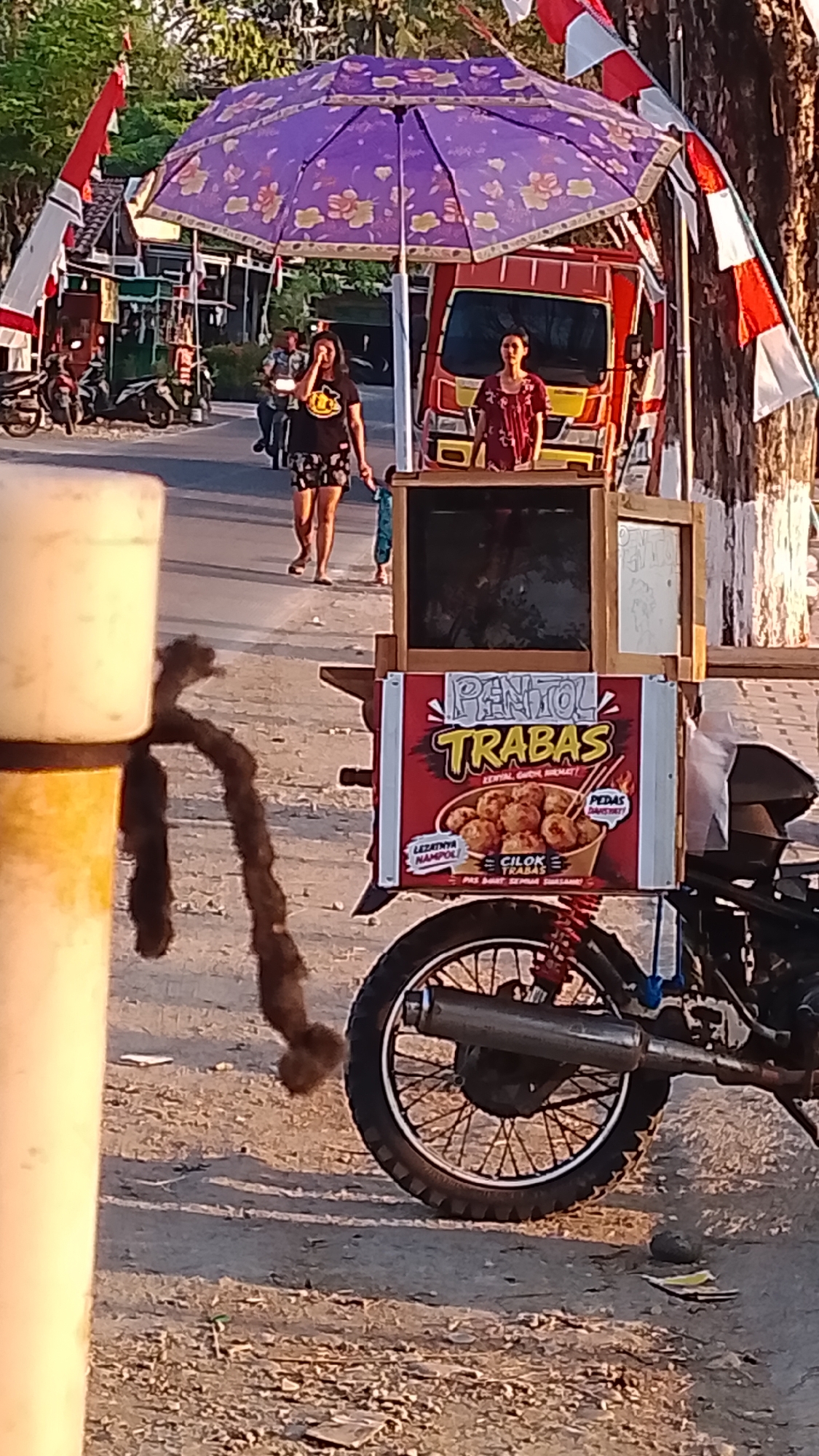 In the city of Karanglangu, Indonesia, a street vendor is operating a food cart. The cart is attached to a motorcycle and is equipped with a large purple umbrella with a floral pattern to provide shade. A red and yellow truck is visible behind the umbrella. Two women are standing near the cart, and one appears to be serving food. A young child is also present. The cart itself is adorned with a sign advertising "PENTOL TRABAS," a type of Indonesian meatball, and depicts a bowl of the food along with text describing its taste and spiciness. In the foreground, a portion of a large, cream-colored cylindrical object, possibly a traffic post or part of the vendor's setup, is visible, along with a dark, rope-like object hanging from it. The ground is covered with gravel and sand, and a tree with red and white Indonesian flags is to the right of the scene. The atmosphere suggests a daytime setting with warm lighting, possibly late afternoon.
