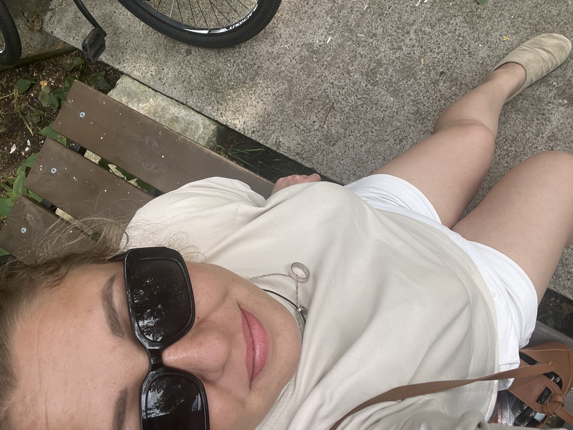 A person is sitting on a park bench outdoors. The person is wearing a light-colored t-shirt, white shorts, sunglasses, and light brown shoes. A small brown purse is hanging from their shoulder. A bicycle is visible to the right of the bench. The ground is concrete and dirt. The environment appears to be a park or public outdoor area. There is no specific information in the image to suggest the location is Chaikovsky, Russia.