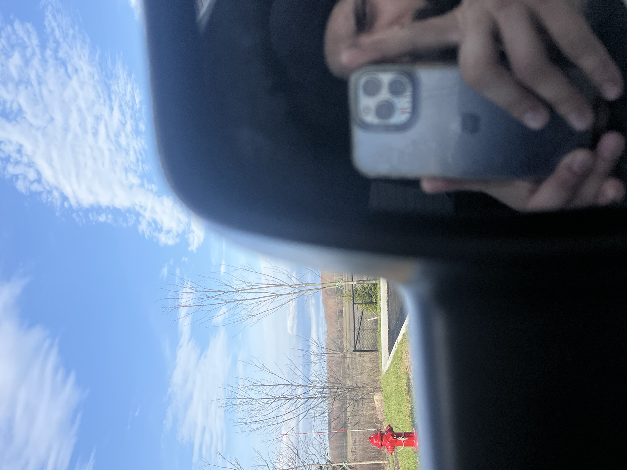 The image shows a selfie taken in a vehicle. A person is holding a cell phone with multiple lenses in their hands. The background shows a clear blue sky with sparse clouds, bare trees, a red fire hydrant, a fence, a road, and a grassy area. Based on the hydrant style and surrounding environment, it is likely in a suburban setting within the Village of Montgomery, United States of America.