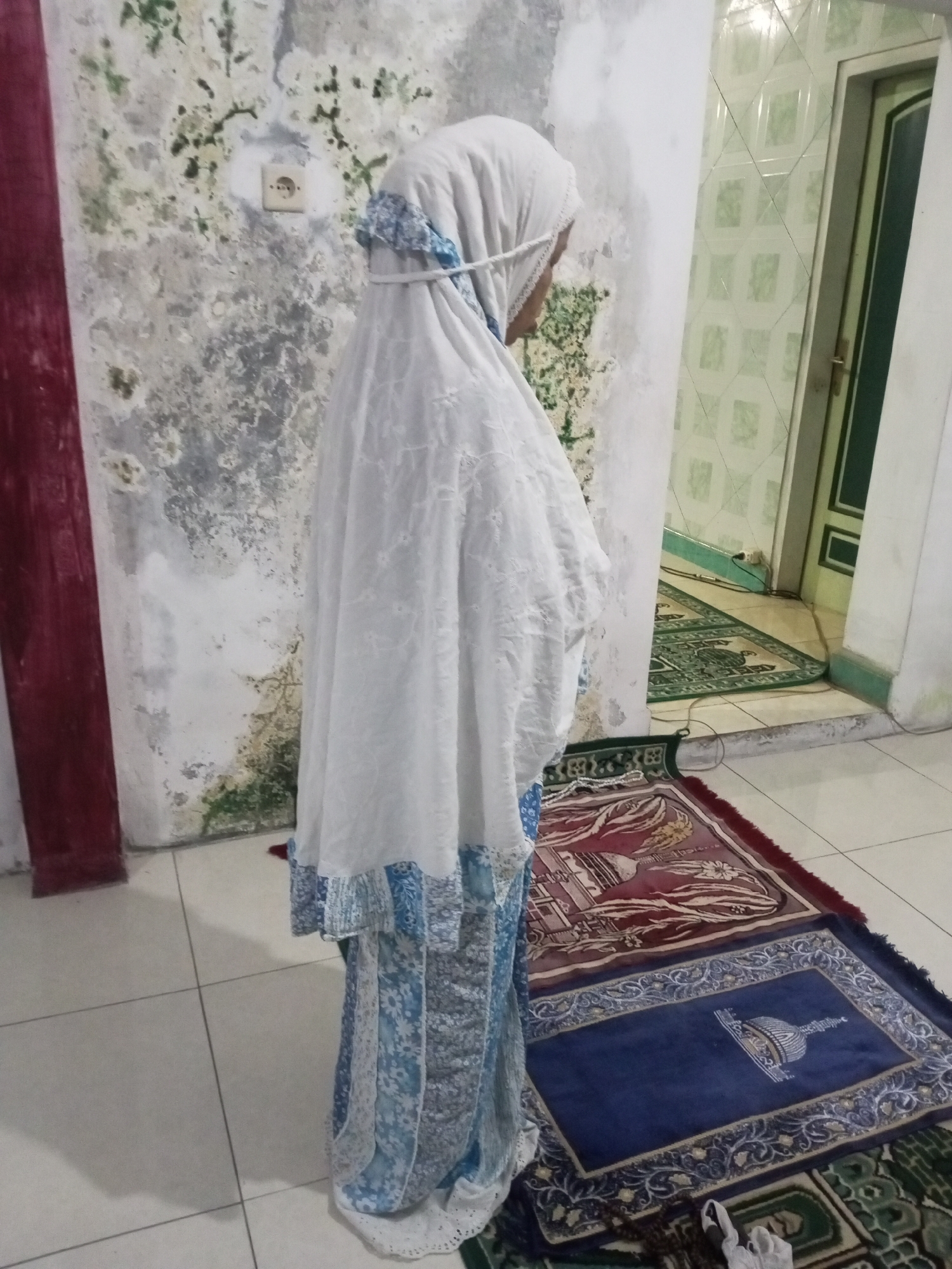 In Andongsili, Indonesia, a person stands on a tiled floor, facing away from the camera, performing what appears to be a prayer. They are covered from head to toe in a white prayer garment, with intricate blue patterns visible towards the bottom. The garment is secured around the head with a white tie. The background reveals a wall with a distressed, textured surface, marked by patches of green and grey. An electrical outlet is visible on the wall. To the right, a door with a dark green frame and paneling is slightly ajar, revealing a glimpse of a room beyond. On the floor, multiple prayer mats are laid out. One is a dark blue mat with a central mosque graphic and ornate borders, placed on top of a larger, maroon-colored prayer mat with floral and architectural motifs. Another green prayer mat is partially visible in the background. The overall scene suggests a domestic or religious space.