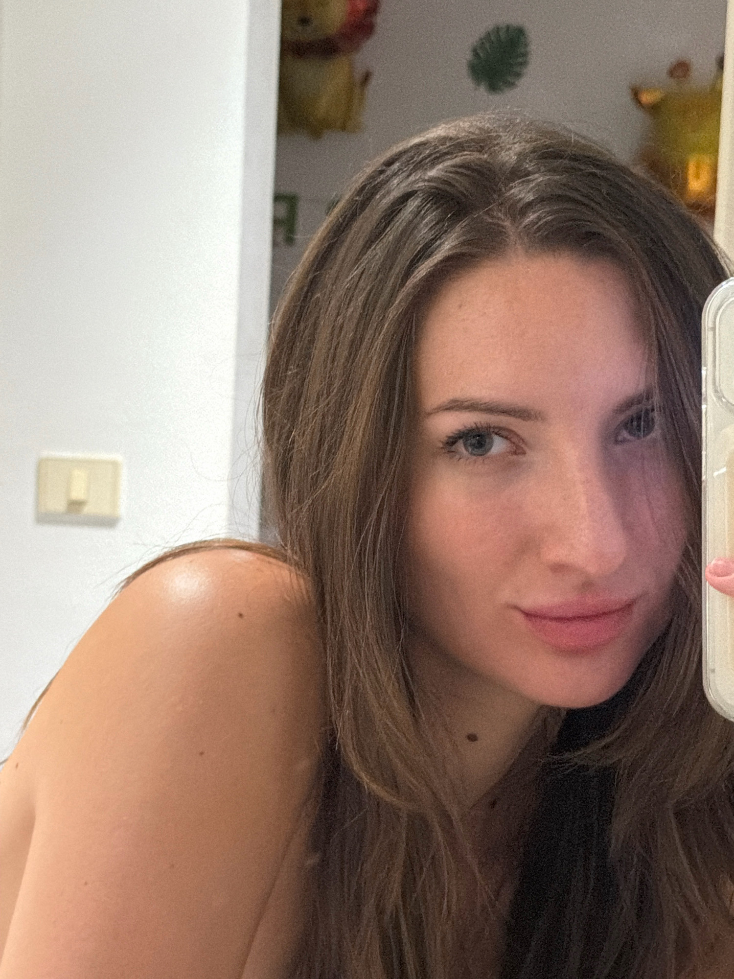 The image is a close-up, selfie-style photograph taken indoors, likely in a residential setting. The subject is a young woman with long, brown hair, looking directly at the camera. She has blue eyes and light pink lipstick. She is wearing a nude-colored top, with her shoulder visible.
To the left of the woman, a white wall is visible with a beige electrical outlet. In the background, out of focus, are some cartoon character decorations, possibly Winnie the Pooh figures, and a green decorative item resembling a leaf. The lighting appears to be artificial, casting soft shadows. The image was taken with a phone, as indicated by the corner of a phone case visible on the right side.
There is no discernible connection to Sharm El Sheikh, Egypt, as the image lacks any specific environmental or cultural markers of that location.