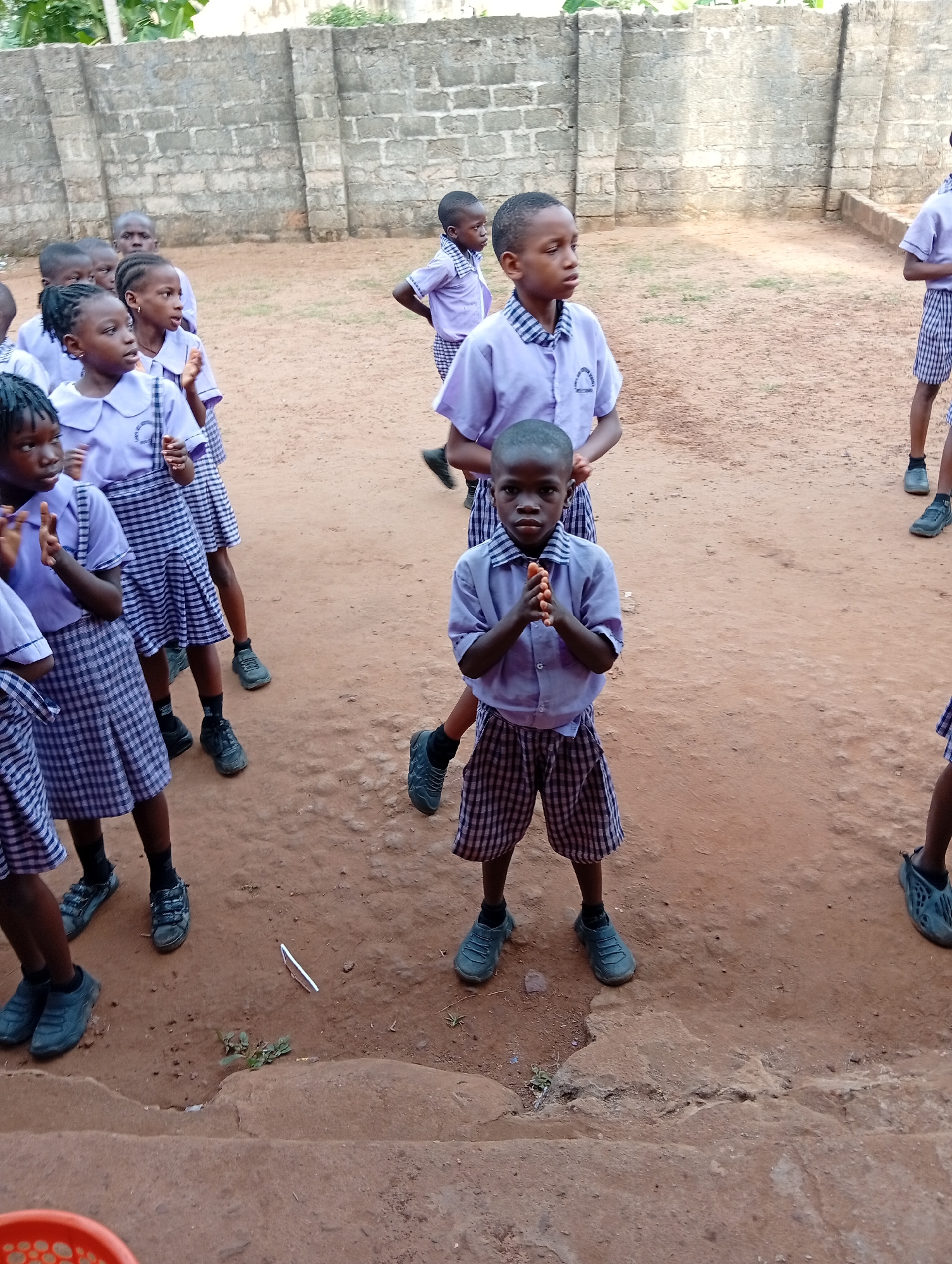 A group of schoolchildren, likely primary or junior secondary students, are standing outdoors on what appears to be reddish-brown earth. The location is likely in Ukwu-Abwa, Nigeria. Most of the children are wearing school uniforms consisting of light purple shirts and blue and white checkered skirts or shorts. They wear dark-colored shoes. Some students have their hands up, and others have their hands clasped in front of them. The background features a weathered concrete wall. The scene suggests a schoolyard or gathering area in a rural or semi-rural setting.
