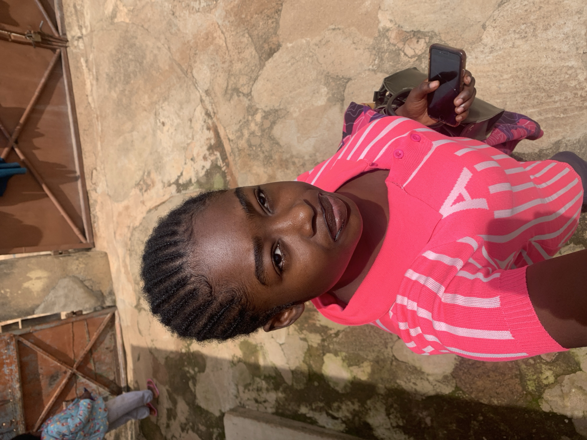 In Rafin Par, Nigeria, an over-the-shoulder shot captures a young Black girl with braided hair. She is looking at the camera, her face angled slightly. She wears a bright pink collared shirt with white stripes. Her left hand is visible, holding a dark-colored smartphone. A dark green bag is slung over her right shoulder, with a patterned cloth draped over it. The background consists of a textured, sandy-colored ground with scattered patches of moss. In the distance, a rusty metal gate and a section of a building are visible. To the left of the frame, a person wearing a light blue, patterned jacket and grey pants, with pink flip-flops, is partially visible walking away.