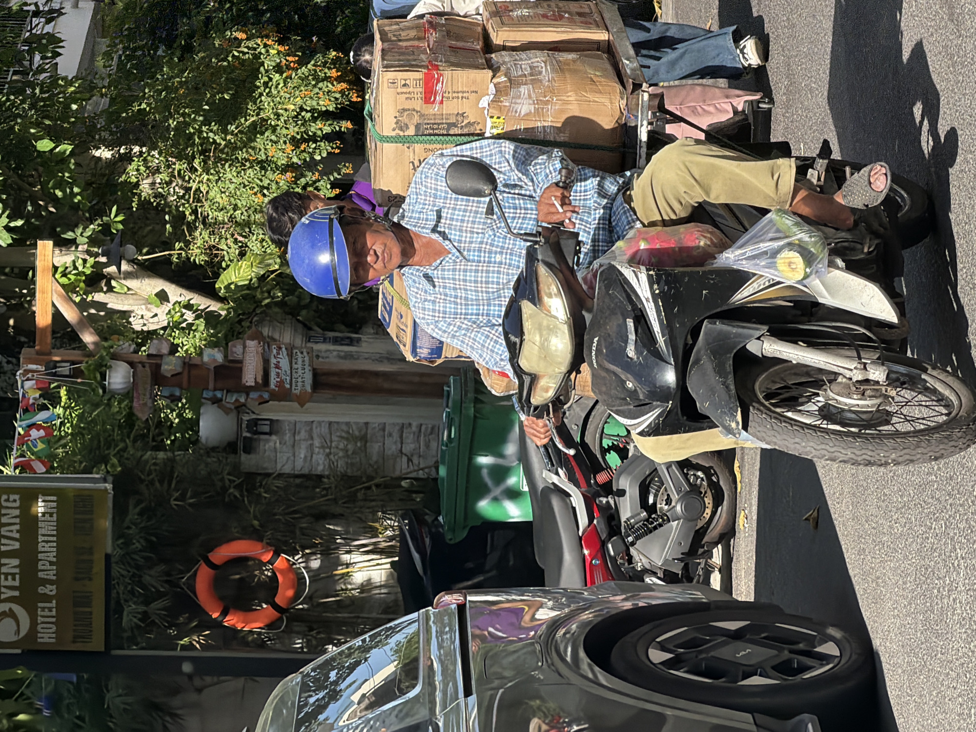 A man wearing a blue helmet and a plaid shirt is riding a scooter loaded with boxes in Nha Trang, Vietnam. The man is holding a cigarette in his right hand. The scooter has a large stack of brown cardboard boxes strapped to the back, with some of the boxes appearing to be sealed with tape. A plastic bag containing fruits and vegetables hangs from the front of the scooter. In the background, there is a sign that reads "YEN VANG HOTEL & APARTMENT". Another scooter is parked nearby, and a car is visible on the left side of the image. The environment appears to be a city street with trees and buildings.