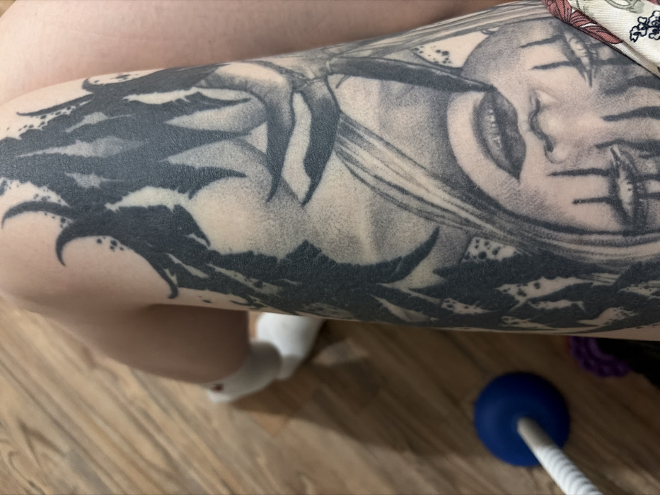 A person's thigh is adorned with a black and grey tattoo depicting a face with eyes closed and markings or tears running down the face. The bottom portion of the tattoo features dark, leafy shapes. Part of a white sock is visible near the knee area. A blue exercise ball and part of a cleaning brush are situated on a wood-grain surface. The person is wearing a flowered patterned fabric at the top of the image.