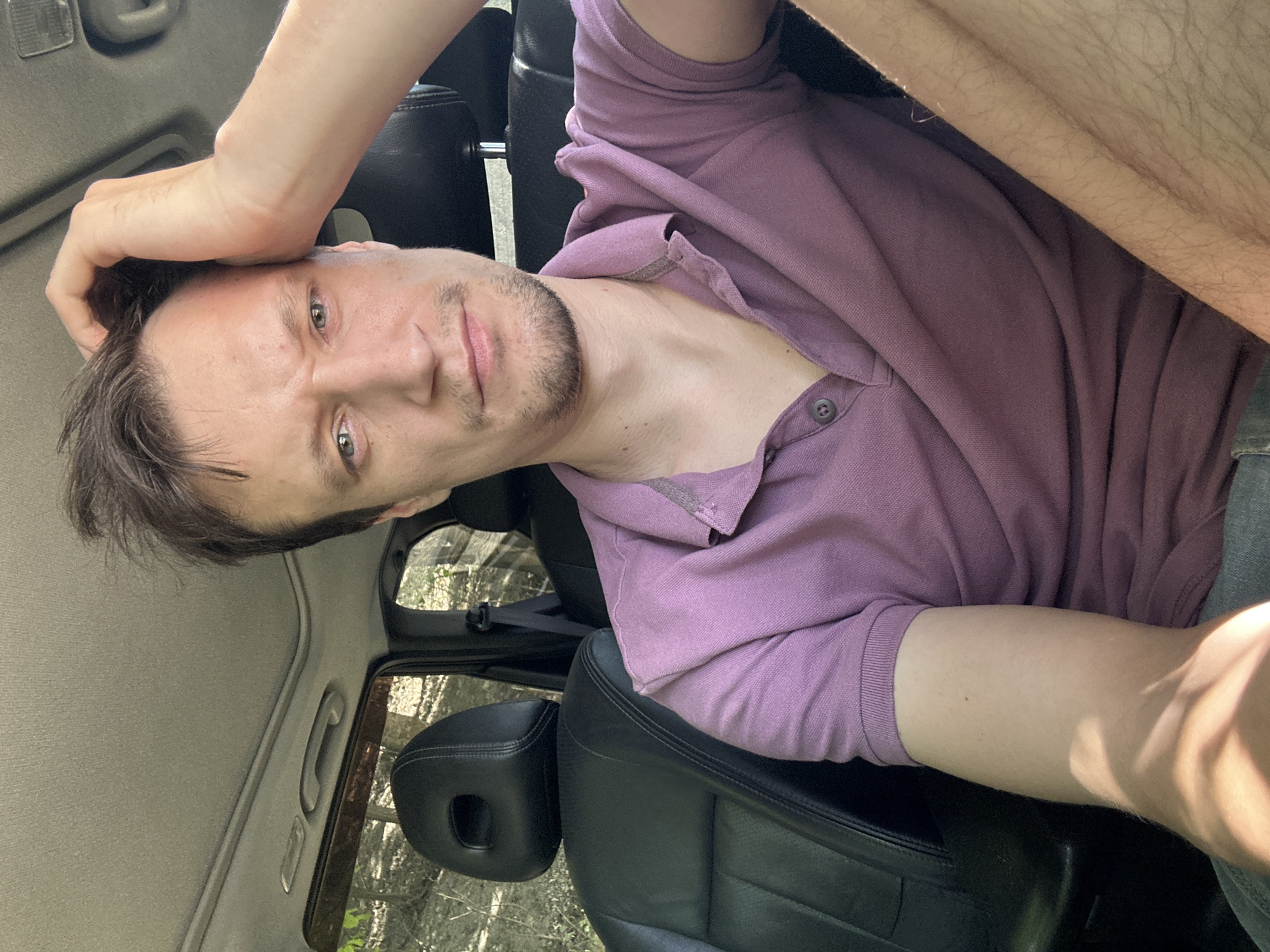 A man is seated in the passenger seat of a vehicle, possibly an SUV, as indicated by the black leather headrest and seat. He has his right hand on his head, appearing to be in thought or perhaps scratching his head. The man is wearing a short-sleeved, collared polo shirt in a muted purple color. Sunlight is filtering into the vehicle, casting shadows on his arm and shirt. The interior of the car includes a sunroof and a glimpse of the ceiling. Outside the car, through the window, trees and foliage suggest a natural, possibly rural setting. Given the context of Makiv, Ukraine, the scene is likely a roadside or a wooded area within or near the village.