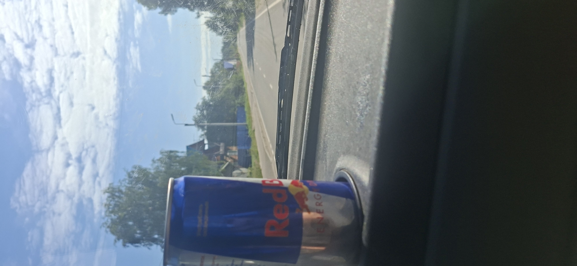 An eye-level medium shot features a can of Red Bull energy drink placed in a car's cup holder, on the dashboard, as seen from the driver's perspective. The can is predominantly blue with the Red Bull logo and a yellow bull graphic visible. The background, visible through the windshield, displays a sunny day with a partly cloudy sky. A road stretches into the distance, lined by trees and houses. The car's windshield wipers are visible across the bottom of the frame. The overall scene depicts a casual moment during a road trip on a clear day in an unspecified rural or suburban location.