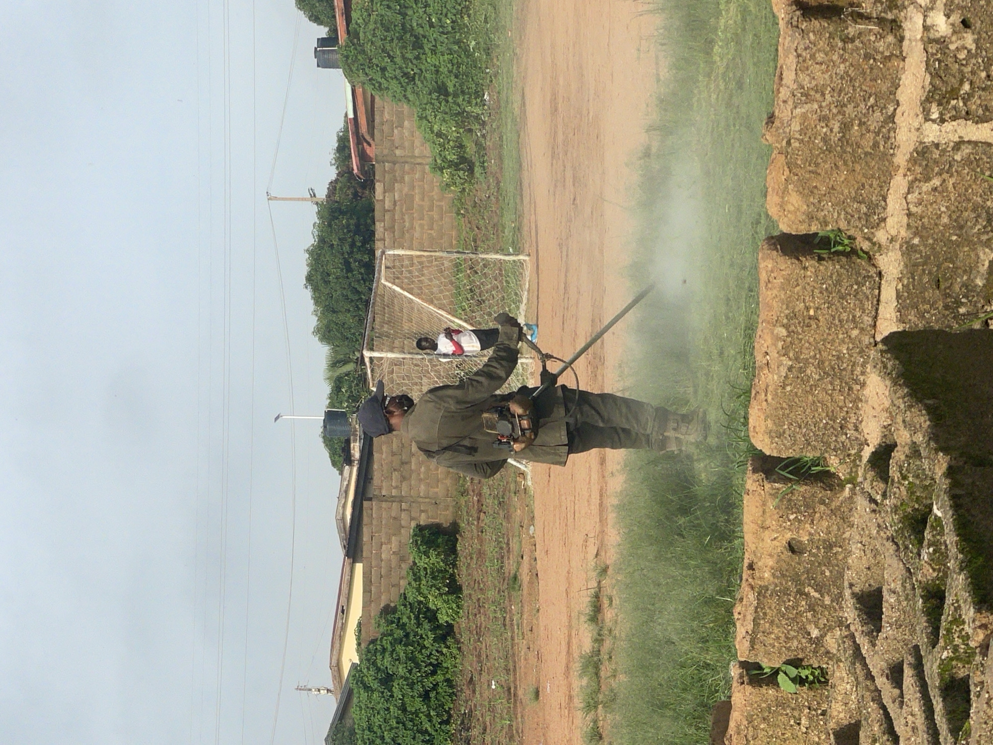A man is using a string trimmer on a grassy area near a dirt soccer field. He is wearing a dark jacket, dark pants, a dark cap, and what appears to be a mask. The string trimmer is emitting a cloud of dust or mist. In the background, a soccer goal is visible, and a young person in a white shirt and red sleeves is standing near it. The area appears to be an urban environment, possibly in Gura Top, Nigeria, with buildings and trees visible behind a brick wall. The ground is a mix of dirt and grass. The sky is overcast.
