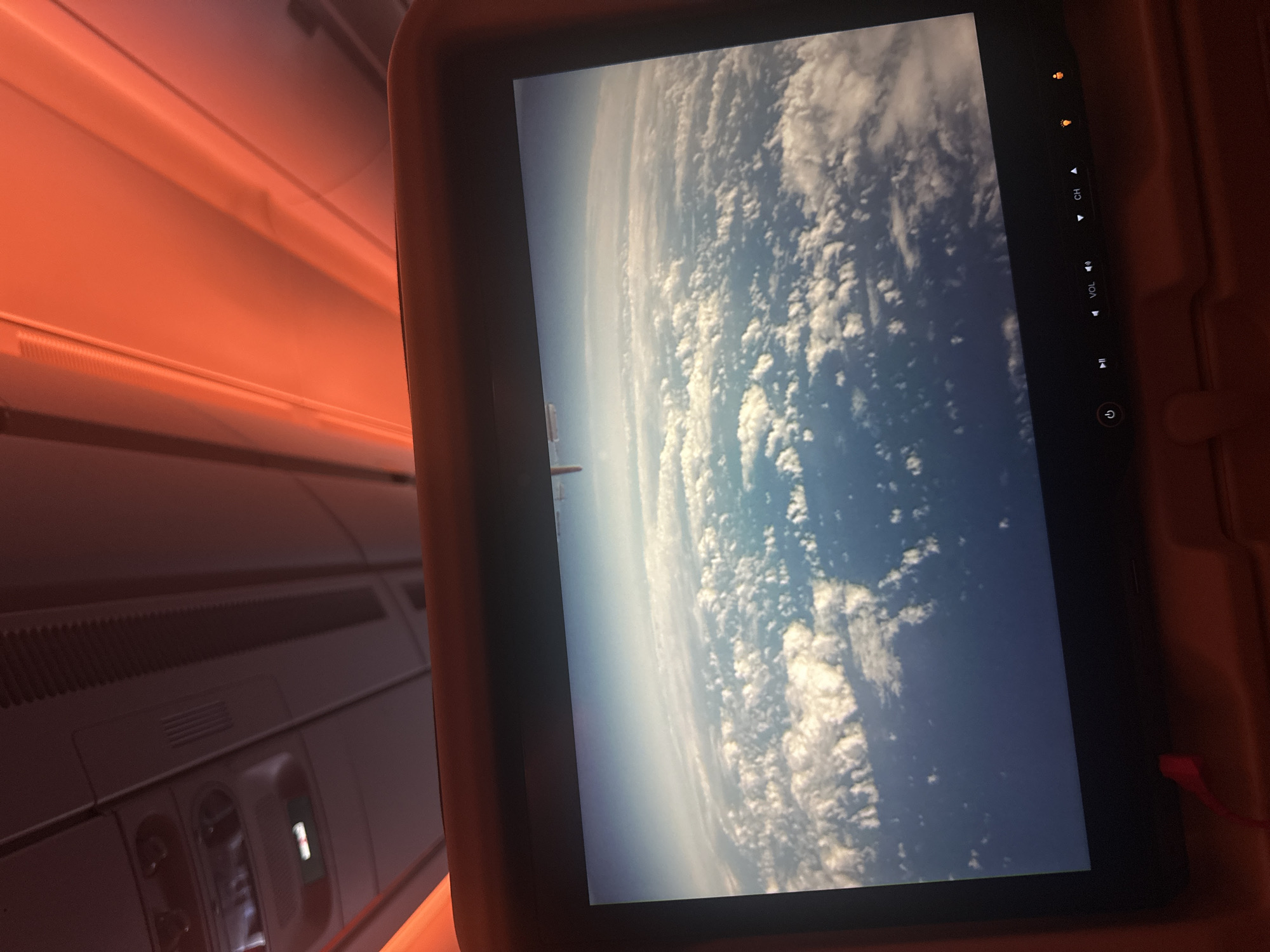 The image shows an airplane cabin interior illuminated with a warm, reddish light. A screen displays a view of Earth from high altitude, showing a blue planet partially covered in clouds. Part of the cabin interior, including overhead panels with vents and reading lights, is visible to the left of the screen. The screen also displays control buttons (power, volume, channel). An earphone cable is visible on the right. The image gives no information about the city of Guarulhos, Brazil.
