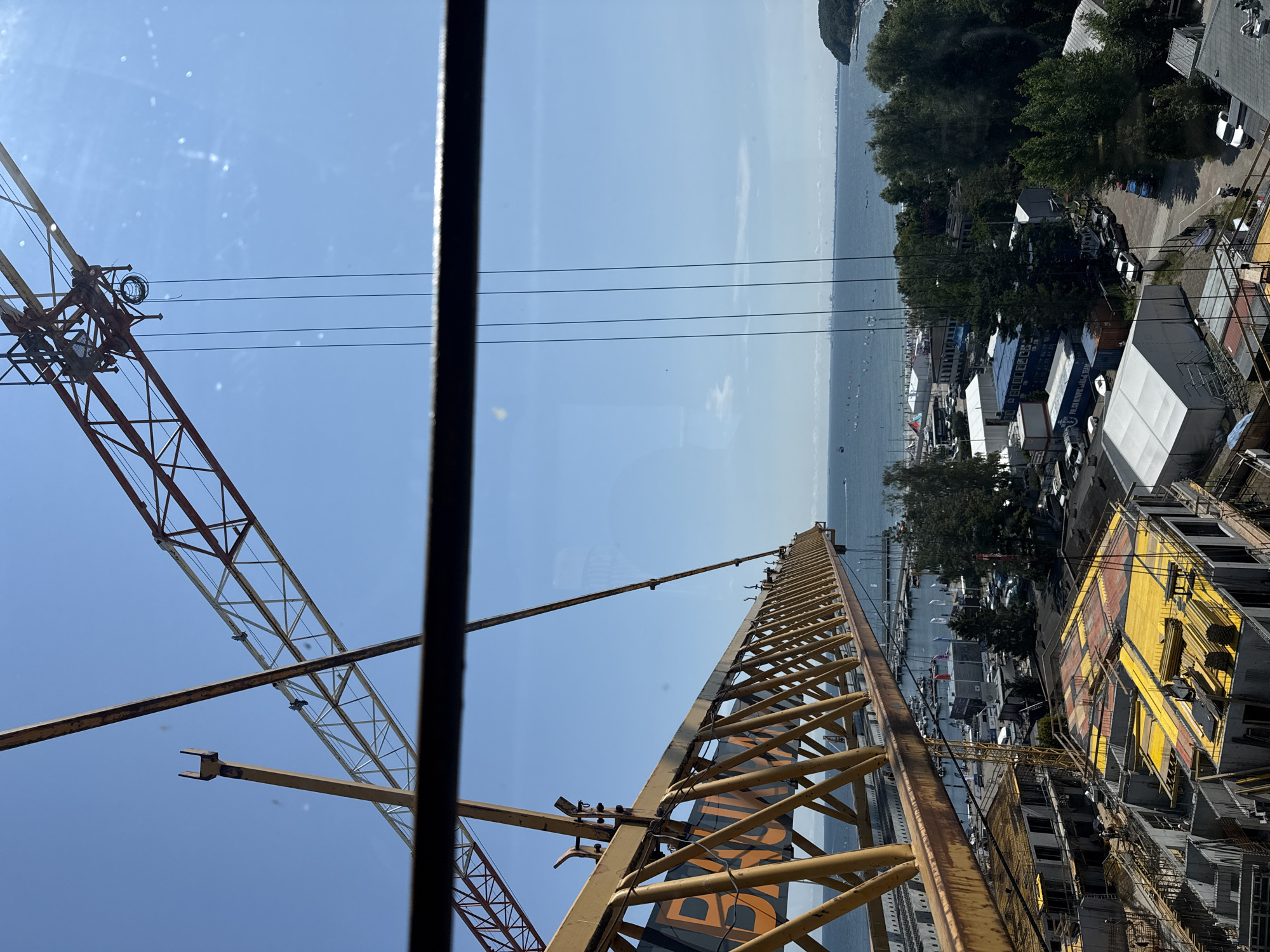 This image captures a construction site with two cranes in Gdynia, Poland, overlooking a bay. The foreground features a yellow construction crane with the word "BUDIMEX" visible on its boom, positioned above a developing multi-story building. The building's structure is partially complete, with concrete frameworks and yellow sheeting covering some sections. In the mid-ground, a marina is visible with numerous sailboats and yachts docked. Beyond the marina, a coastline with trees and buildings stretches across the horizon. The sky is clear and blue. The image appears to have been taken from an elevated position, possibly another construction crane or a high-rise building.
