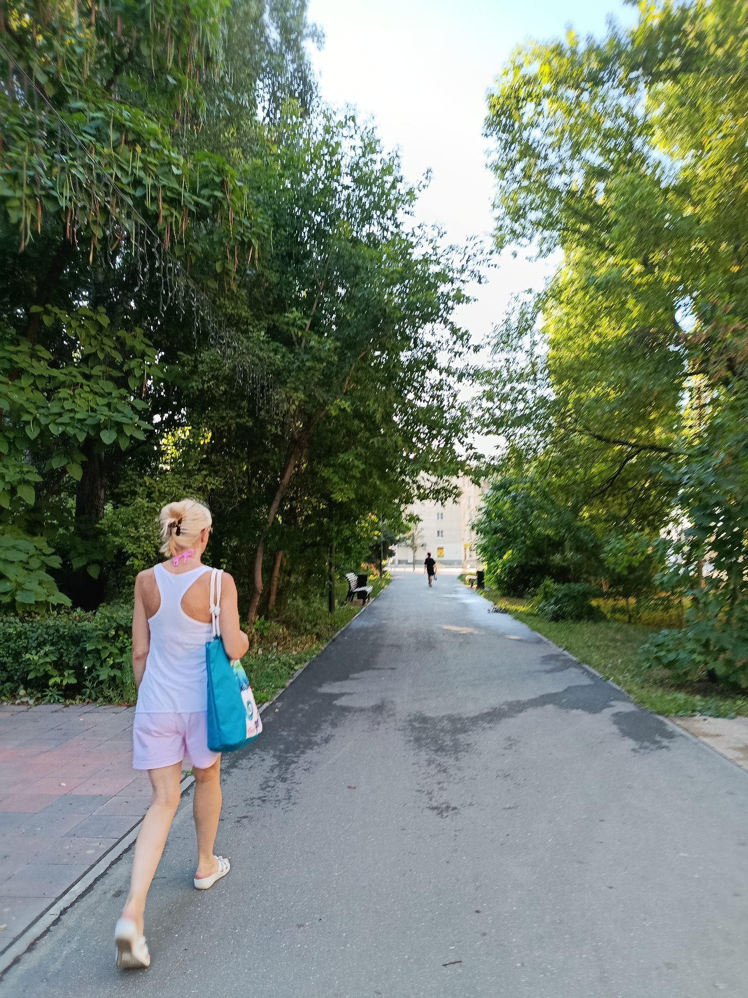 A woman with blonde hair, wearing a white tank top and pink shorts, walks down a paved pathway lined with trees. She carries a blue tote bag over her left shoulder. Further down the path, a person in a dark shirt and shorts is jogging. Buildings are visible in the background beyond the trees. The scene appears to be a park or a tree-lined street in Saratov, Russia.
