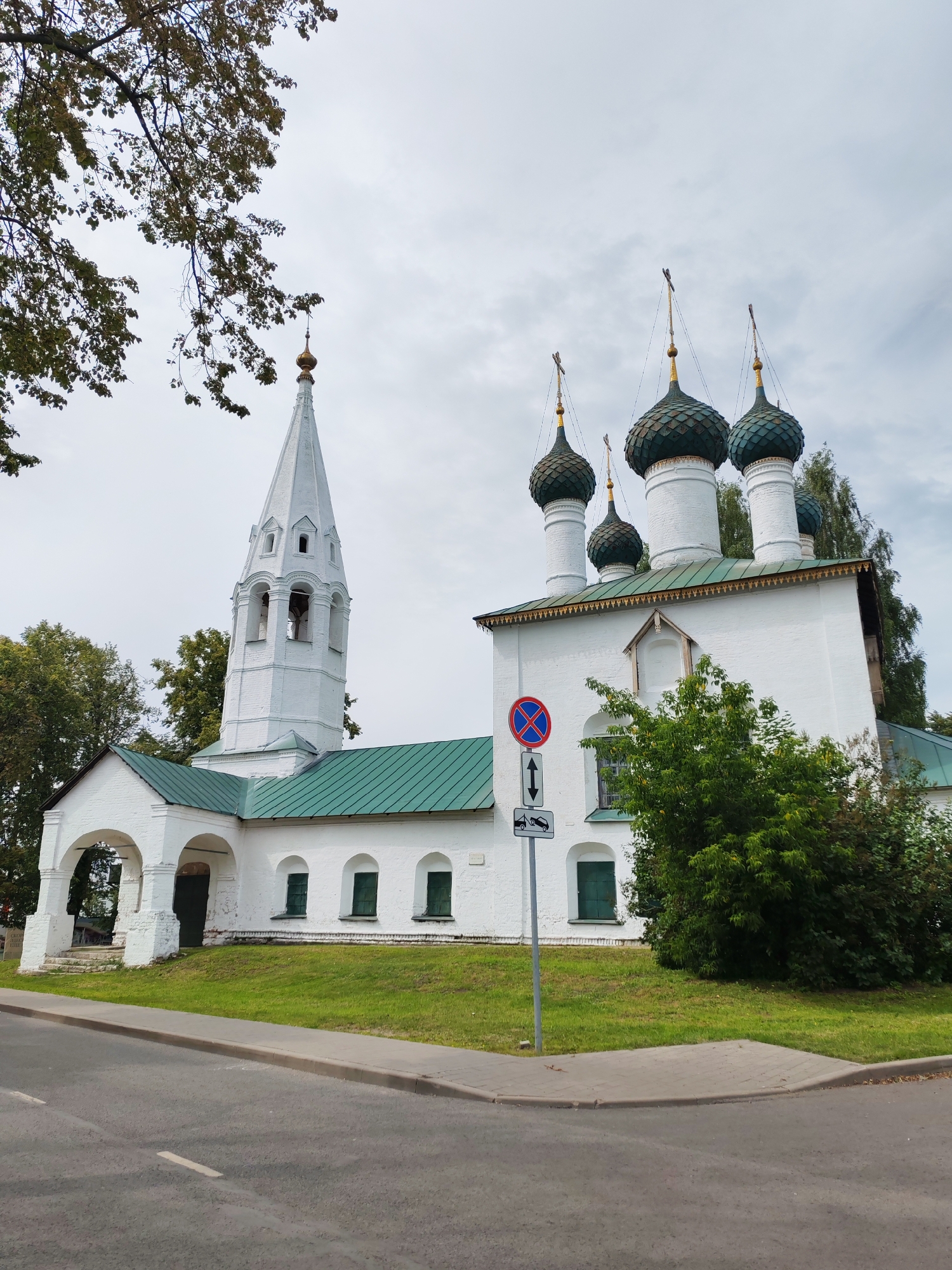 A white, multi-domed Russian Orthodox church with a tall, white bell tower stands under an overcast sky in Rusanovo, Russia. The church has a green metal roof and several green-domed cupolas topped with gold crosses. The bell tower is slender and tapers to a point, also topped with a gold cross. An arched entryway leads into the church. The building is situated on a grassy slope next to a paved road. A traffic sign showing a no-stopping symbol and a tow-away warning is positioned in front of the church. Trees with green foliage partially obscure the view of the church and bell tower on the left side of the image.