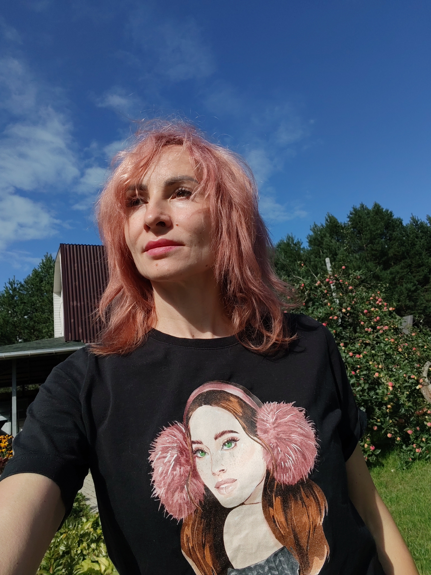 An outdoor, medium shot captures a woman with shoulder-length, pink-tinted hair and fair skin looking to her right, her head tilted slightly upwards. She is wearing a black t-shirt with a large graphic of a woman's face adorned with pink earmuffs. Behind her, a brown-roofed building with white siding is visible, partially obscured by dark green coniferous trees. To the right of the building, an apple tree laden with ripe red apples stands in a grassy area. The sky above is a vibrant blue, dotted with a few wispy white clouds. The scene appears to be a private residence's backyard, possibly in Chaikovsky, Russia, suggested by the presence of a typical rural Russian dacha-style building and apple orchard.