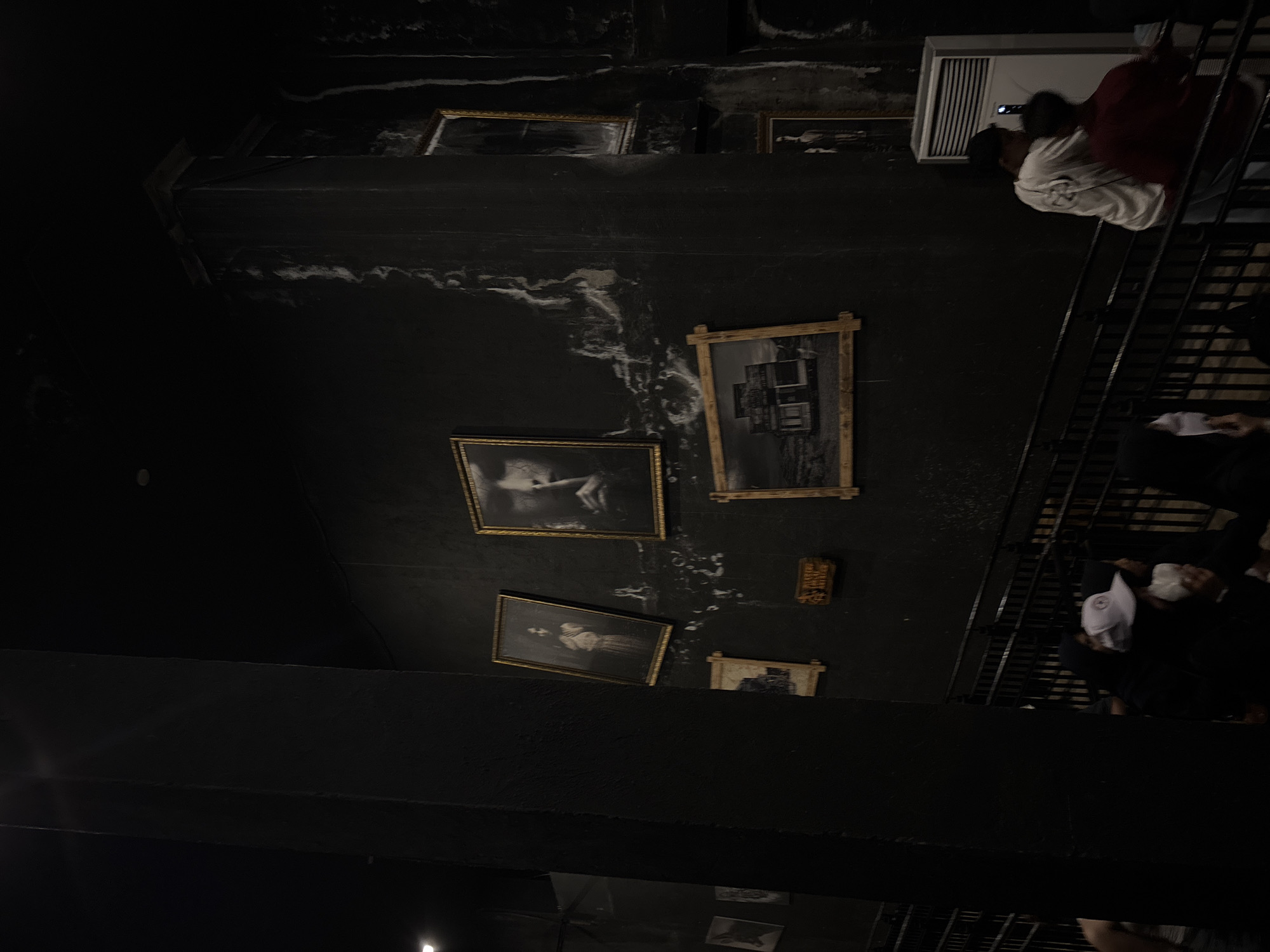 This media file captures a dimly lit, narrow interior space, likely a hallway or staircase, in North Jakarta, Indonesia. The walls are dark, possibly black, featuring striking white, wavy patterns that suggest distressed paint, water stains, or a decorative effect.
Several framed pictures or artworks, set in golden or dark wooden frames, are mounted on the dark wall. These appear to be black and white or monochrome images, including portraits of individuals (one notably a woman in historical attire), a landscape, and abstract compositions, contributing to an antique or somber aesthetic. A small, dark wooden plaque with faint, illegible markings is also visible on the wall.
On the right side of the image, a black metal staircase with railings descends, and several people are present on or near it. High on the wall, a white, rectangular air conditioning unit is mounted. Two individuals are near this unit: one in a white shirt and dark pants, and another in a red top, both facing away from the camera. Further down the stairs, more people are visible, with one person wearing a white baseball cap.
The scene suggests visitors navigating or observing displays within a unique environment, possibly a museum, art gallery, or a themed immersive experience designed to evoke an aged, mysterious, or slightly eerie ambiance. The lighting is low, creating a dramatic and atmospheric mood. No discernible text is clearly legible in the image, other than indistinct features on the wooden plaque and the air conditioner.